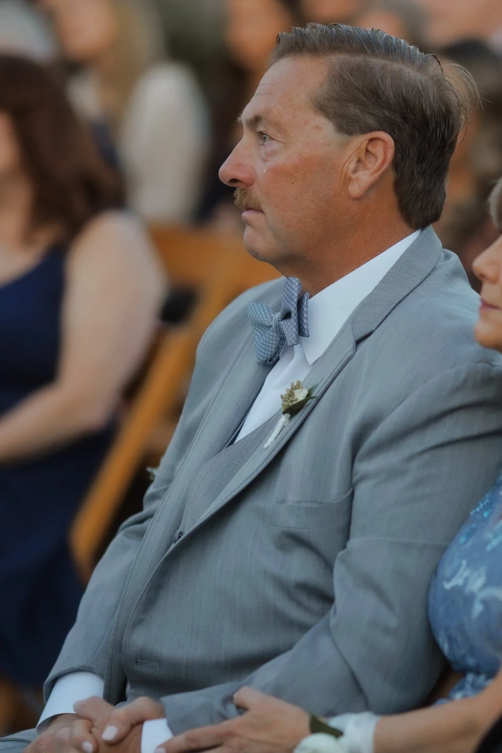 Man in a gray suit and blue bow tie watching the ceremony with a tear on his cheek.