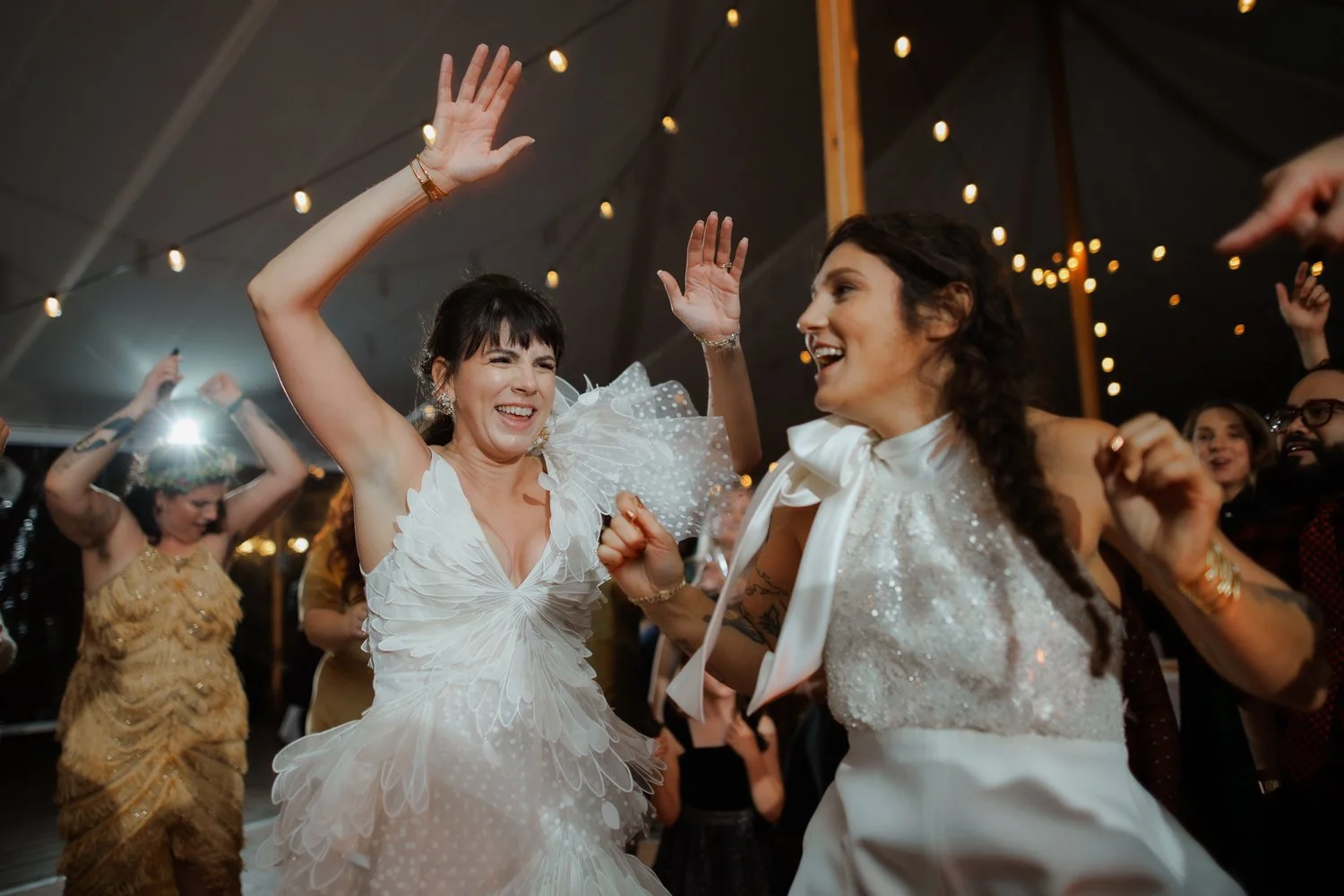 Two brides dancing with their arms raised and big smiles beneath string lights in a reception tent.