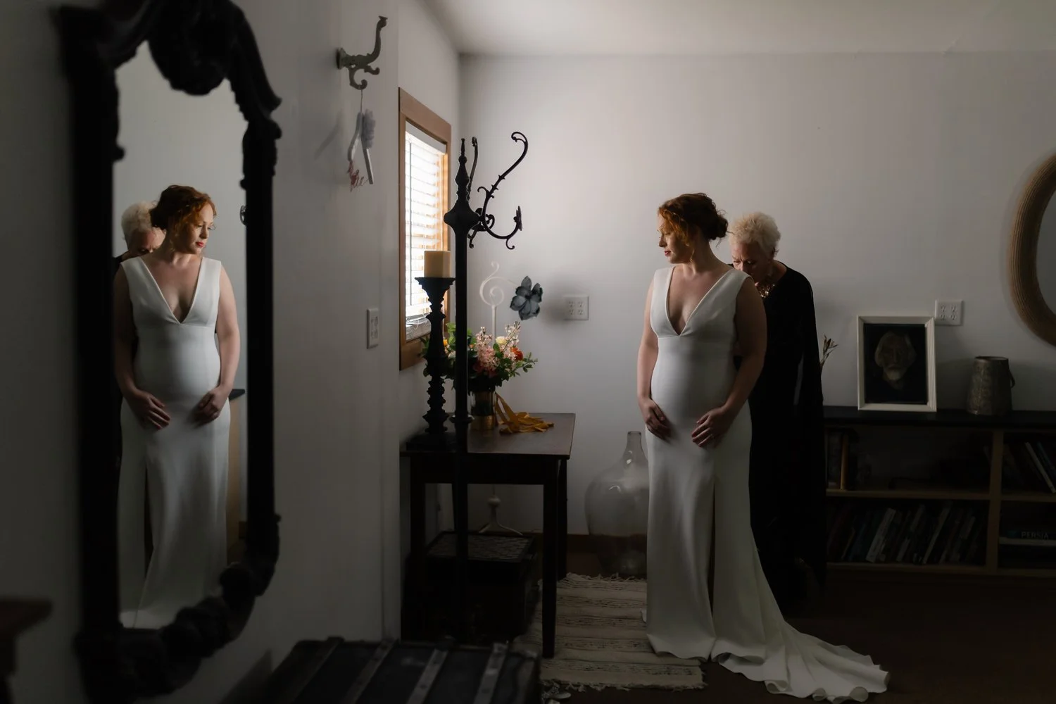 Mom zips up bride's dress with her reflection in the mirror.