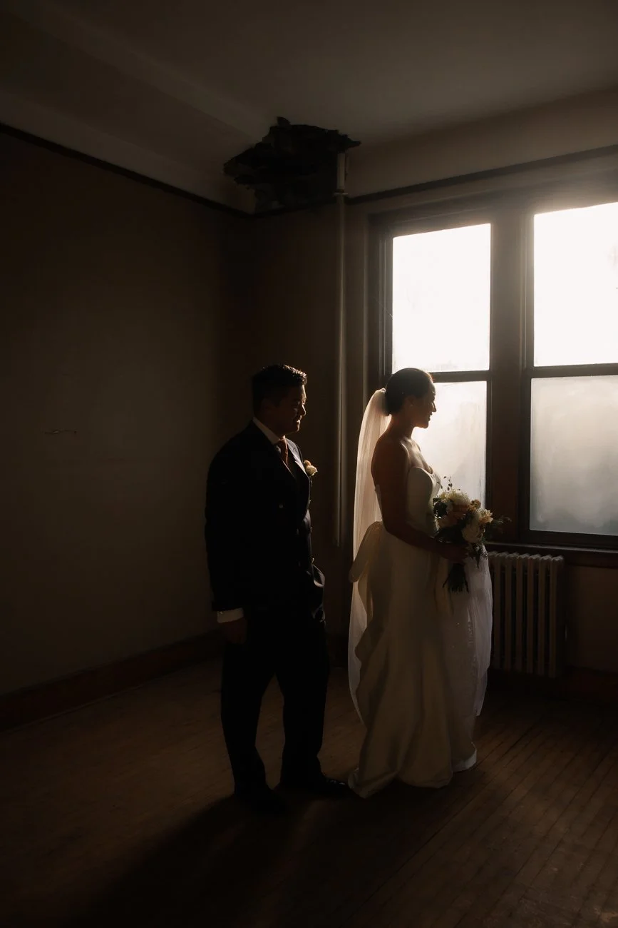Silhouette of a bride holding a bouquet and a groom standing next to her in front of a large window with sunlight shining through, in a dimly lit room.