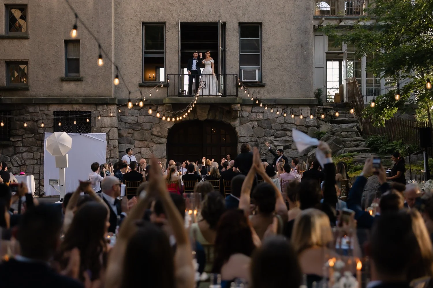 Couple stands on porch overlooking their wedding reception as their guest cheer them on.