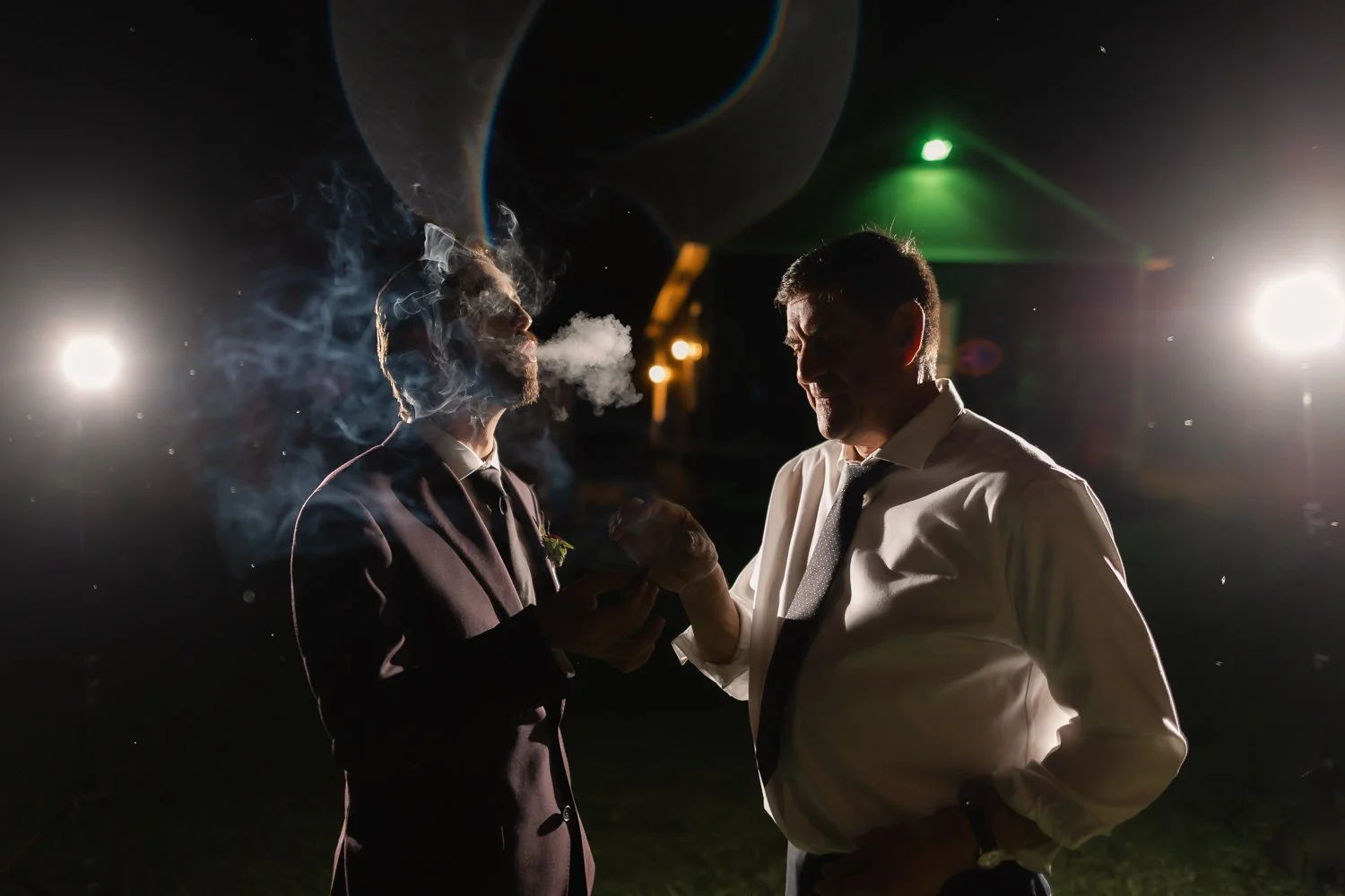 Groom shares a cigar with his dad under night lights at wedding reception.