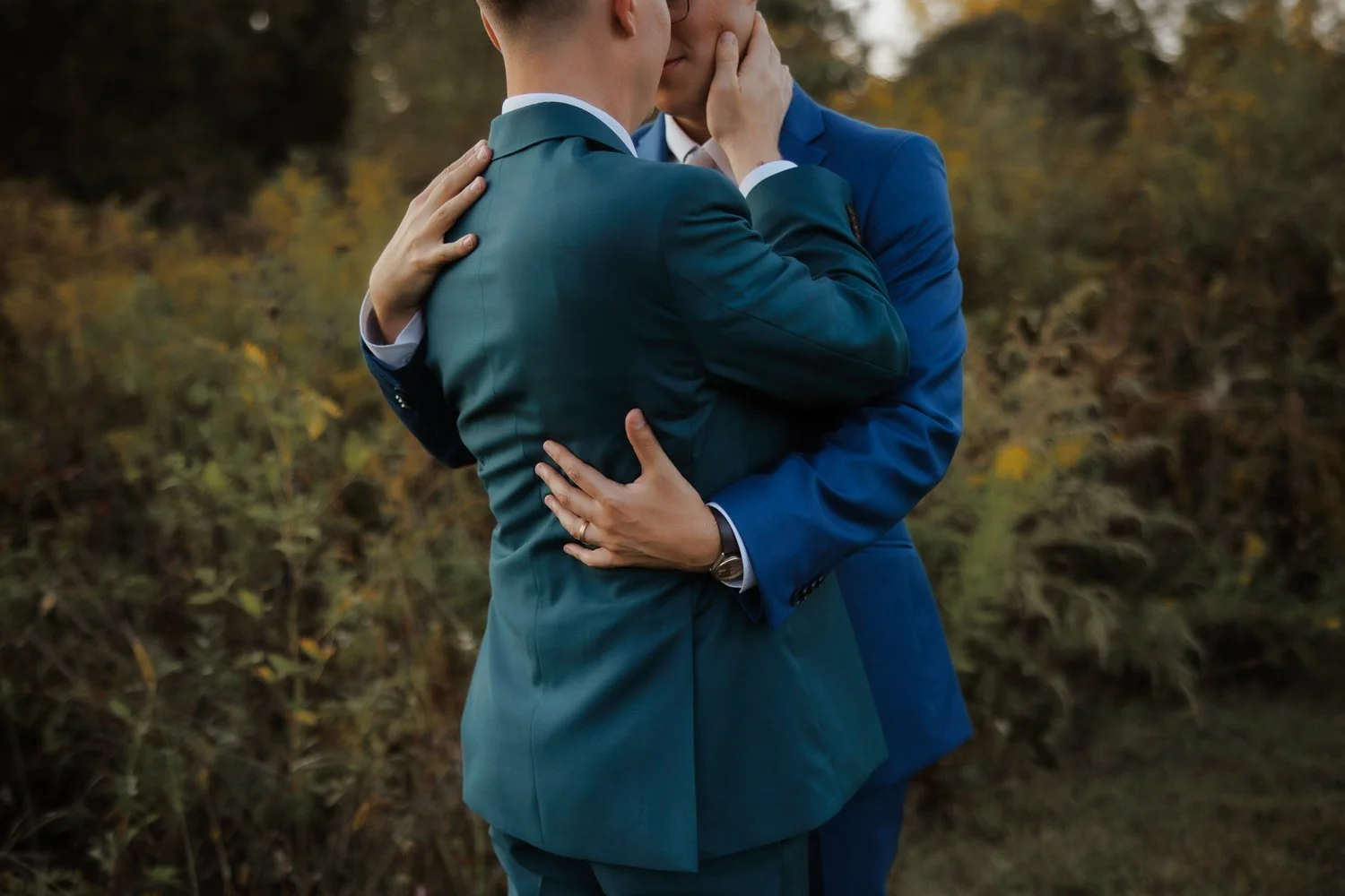 Two grooms in teal and blue suits holding each other close among golden autumn foliage.
