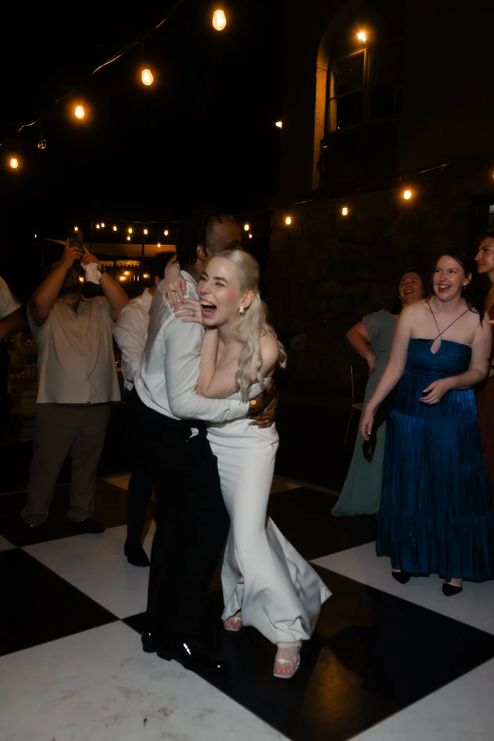 Groom holds onto bride while she laughs over his shoulder on checkered dance floor.