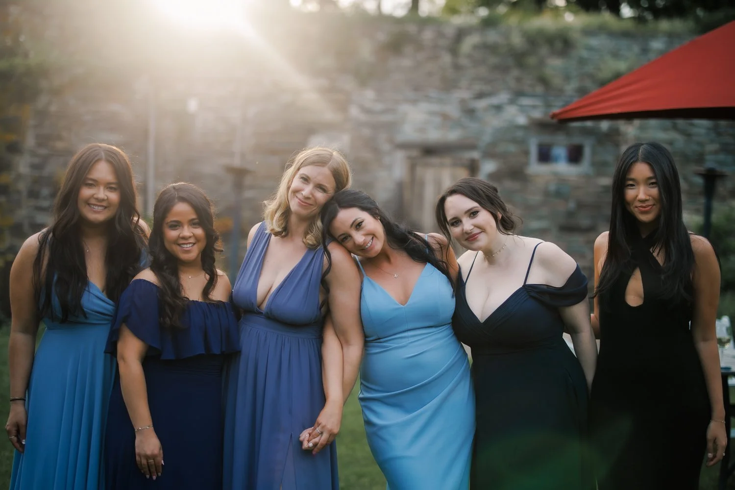 Six bridesmaids in blue and black dresses leaning together and smiling in warm backlight beside a stone wall.