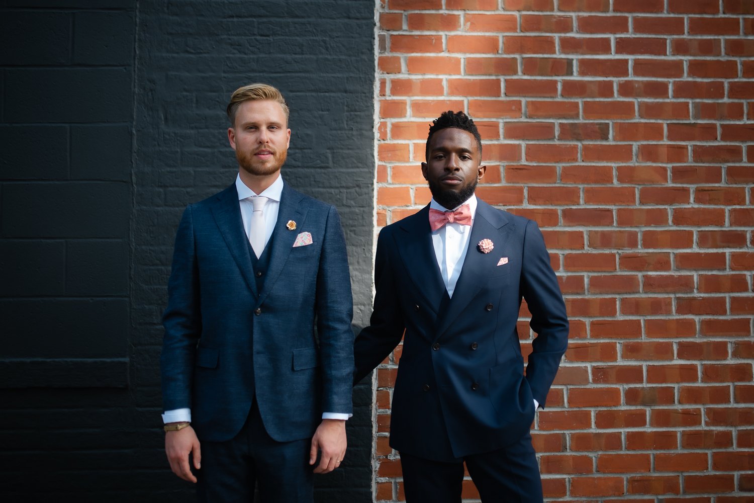 2 grooms stand against contrasting brick walls for editorial portrait.