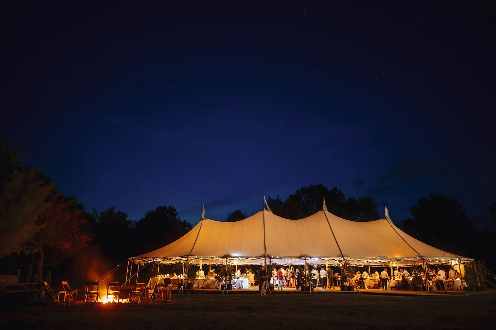 Illuminated reception tent glowing at dusk with a bonfire burning nearby.