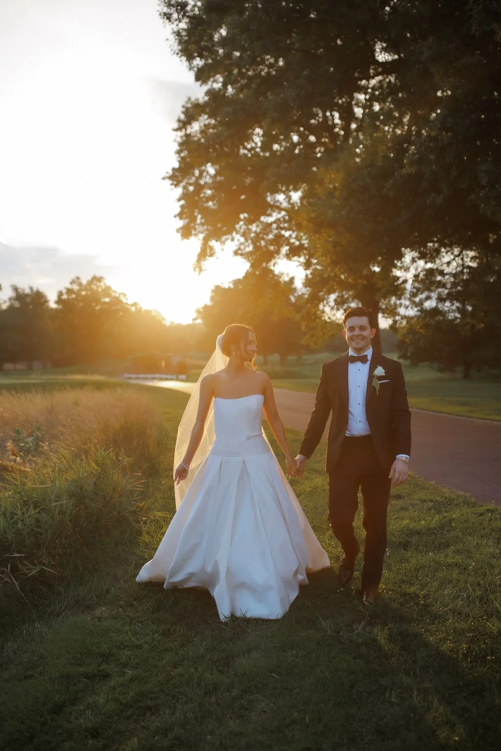 A bride and groom holding hands, walking outdoors during sunset, with large trees and a grassy area in the background.