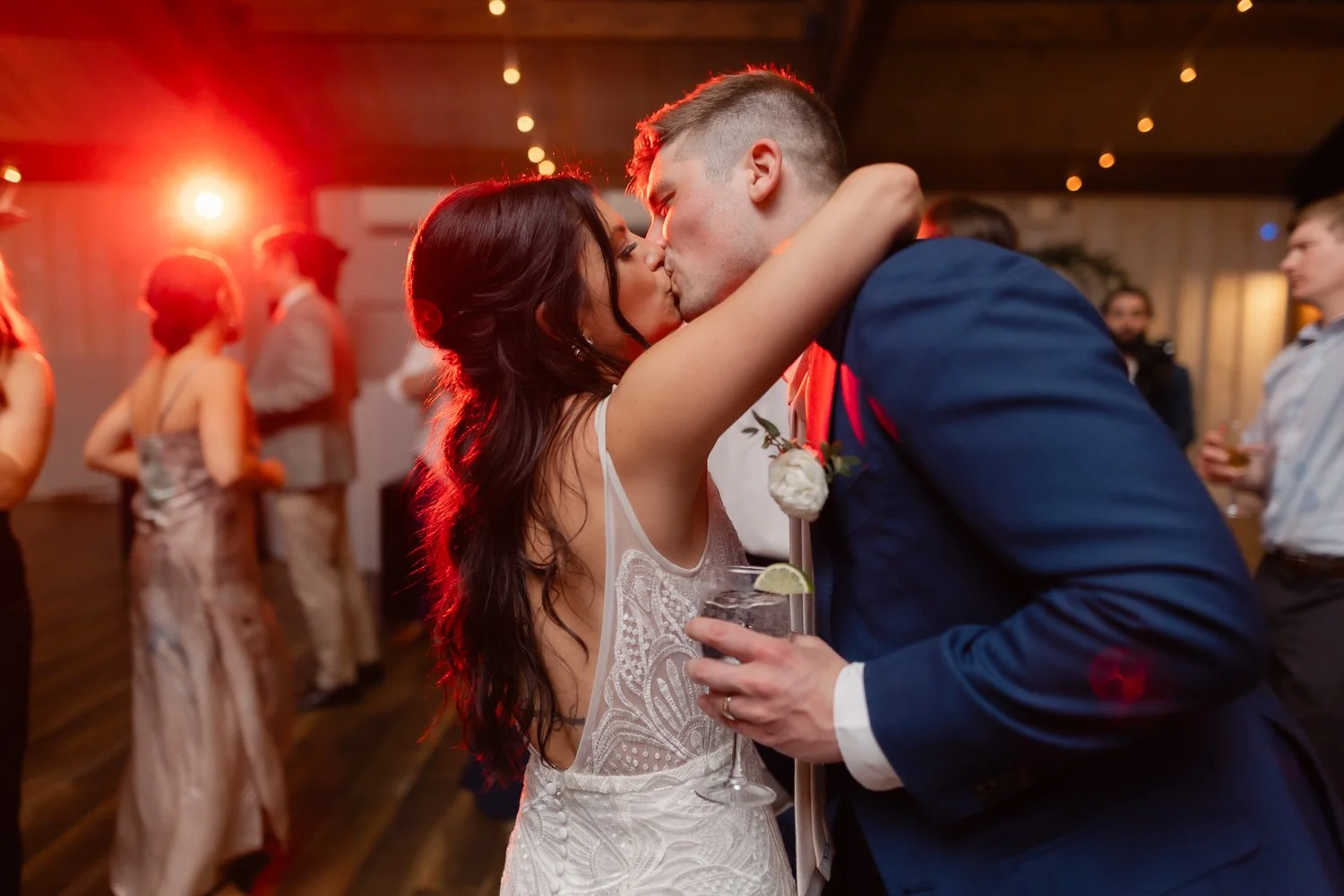 Wedding couple kisses under red party lights on dance floor.