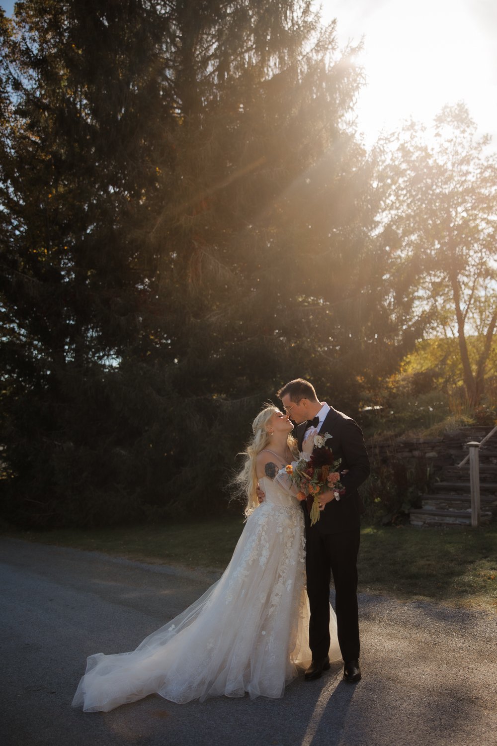A bride and groom sharing a kiss outdoors on a sunny day, with the bride in a white wedding gown and the groom in a black tuxedo holding a bouquet of flowers.