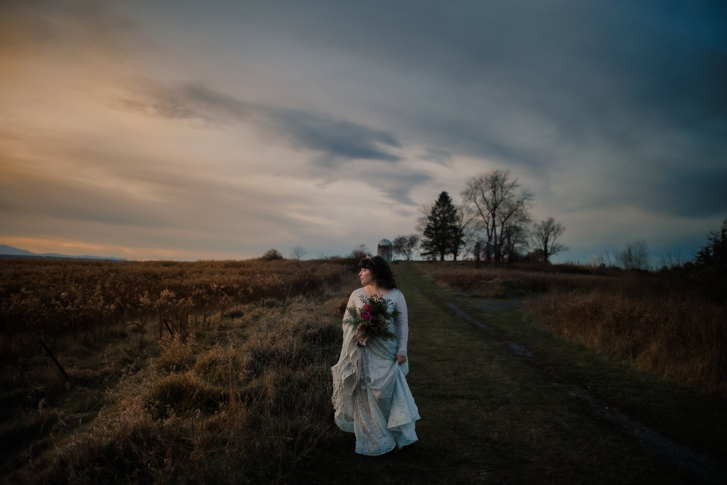 Bride walks down path in huge field with moody cloudy sky behind her.