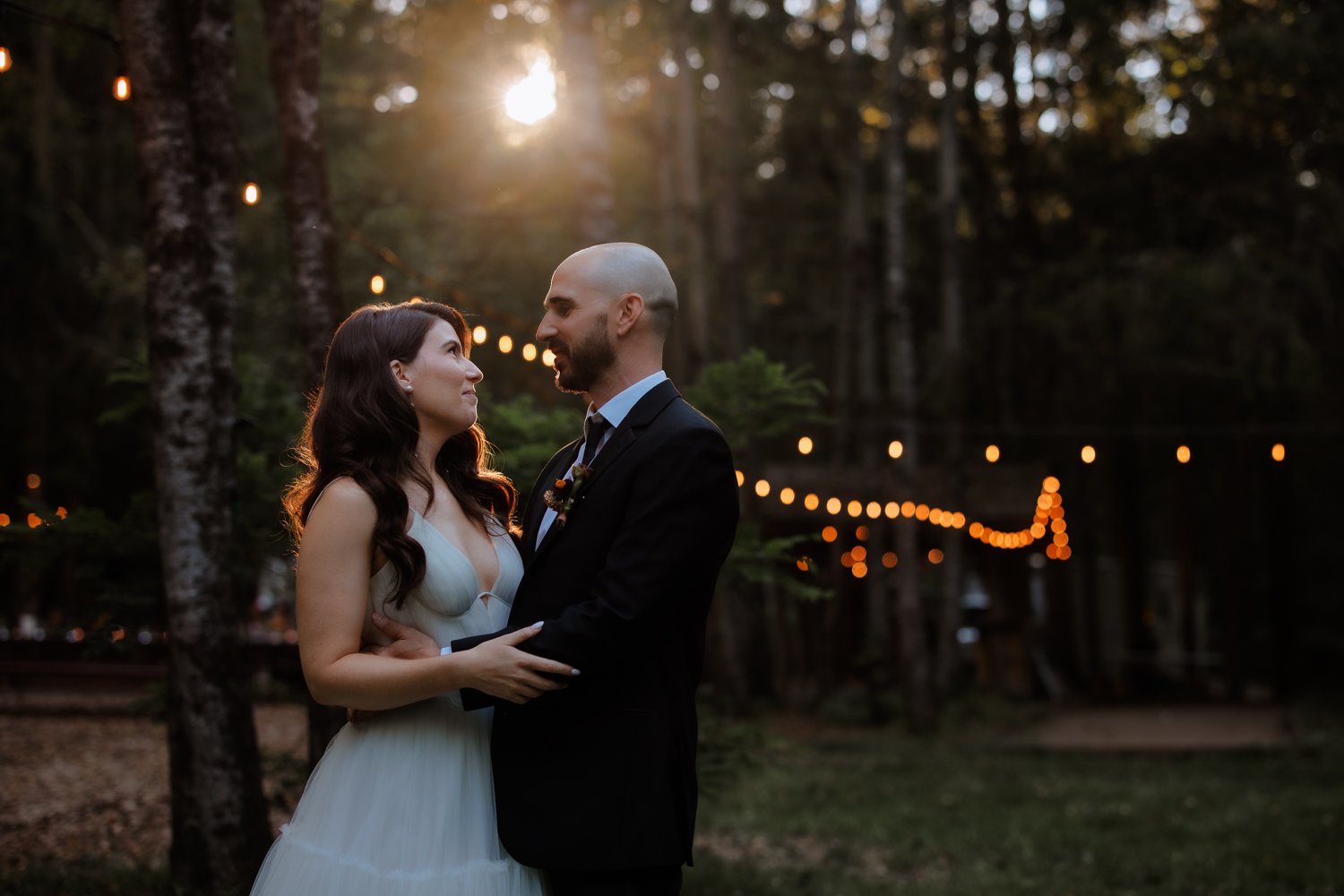 A couple in formal attire embracing outdoors with string lights and a forest background, during sunset.