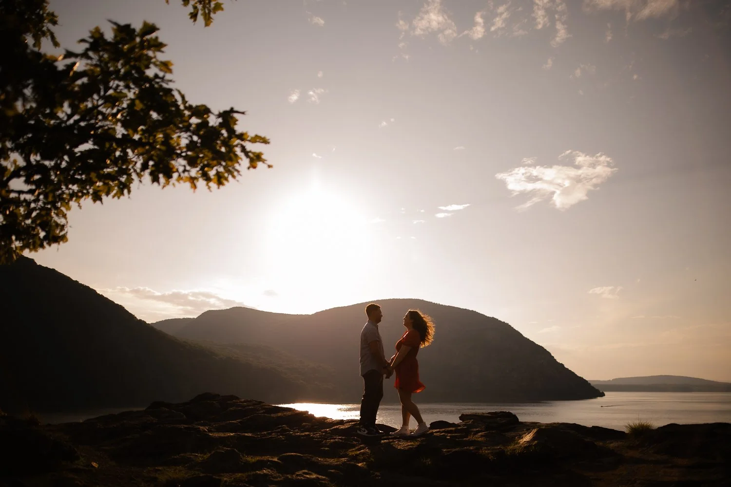 A couple holding hands and facing each other at sunset by a lake with mountains in the background.