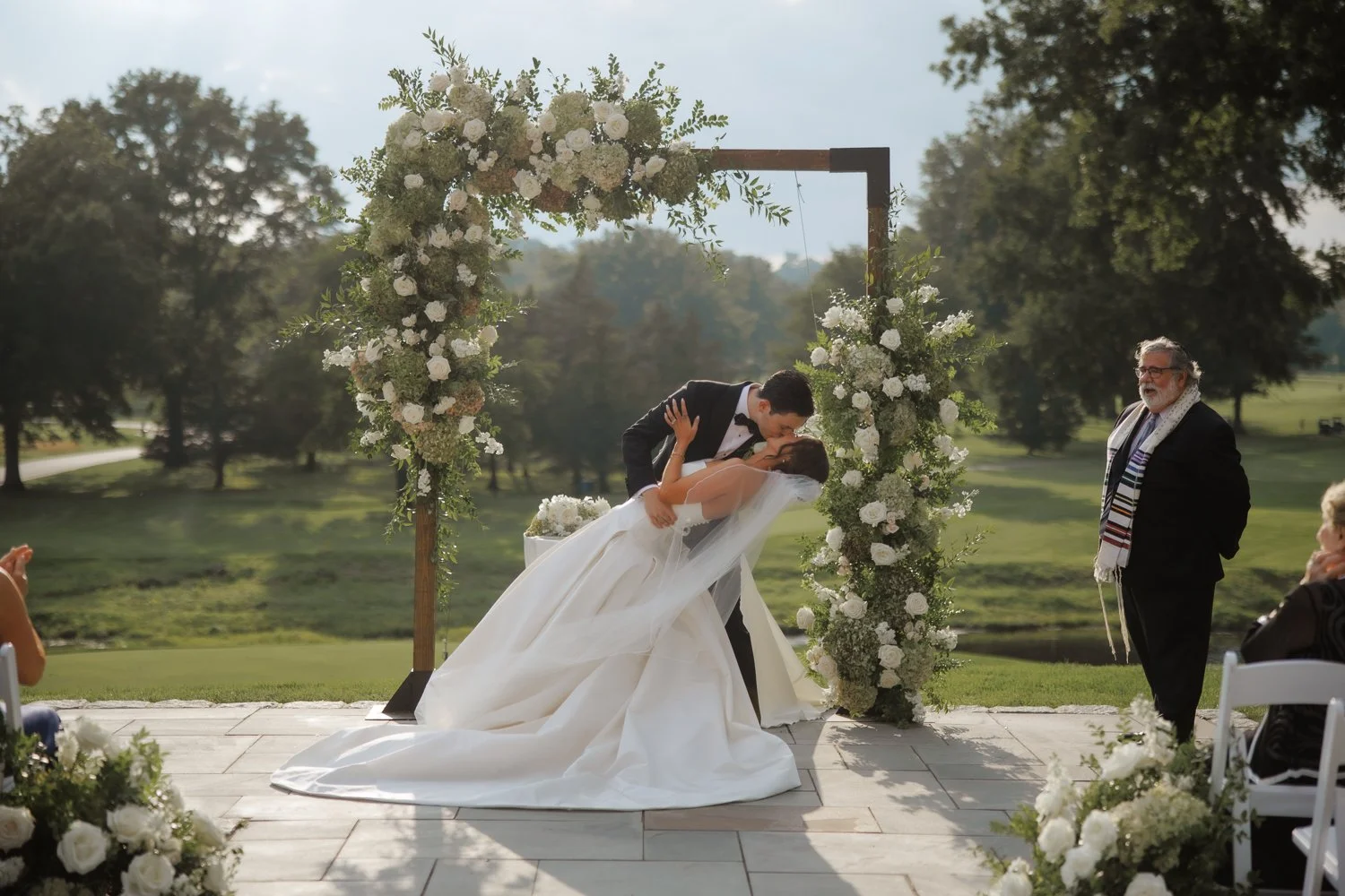 A bride and groom kissing under a floral wedding arch outdoors during their wedding ceremony, with officiant and guests watching.