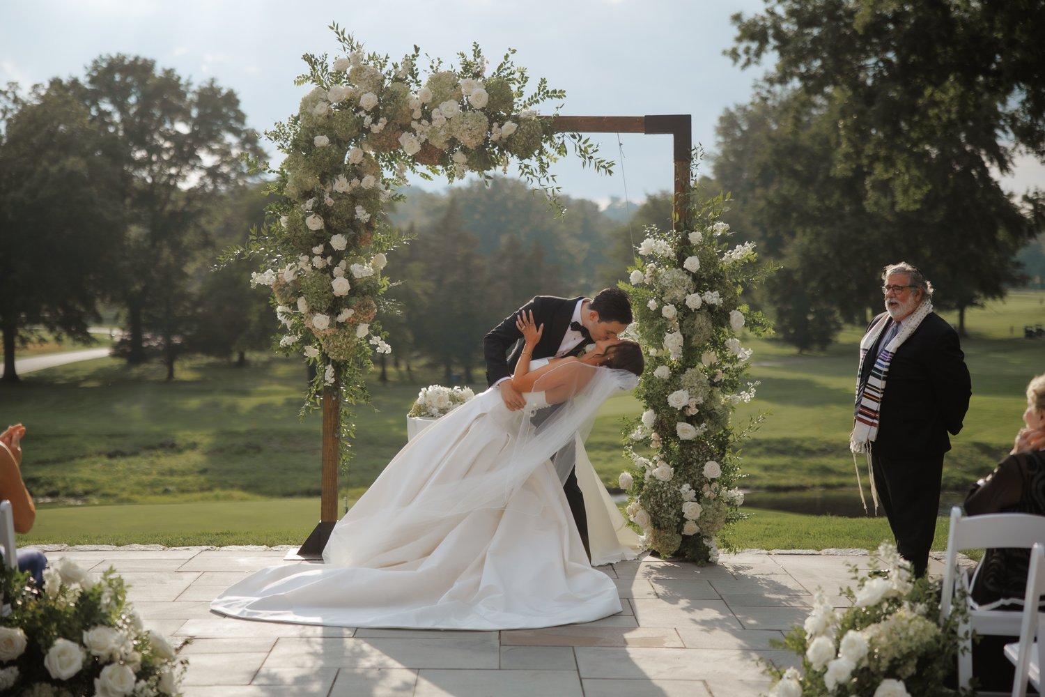 A bride and groom share a kiss during their outdoor wedding ceremony beneath a floral arch. The bride wears a white wedding gown and veil, and the groom wears a black tuxedo. An officiant stands nearby, watching the couple, with trees and a sunny landscape in the background.