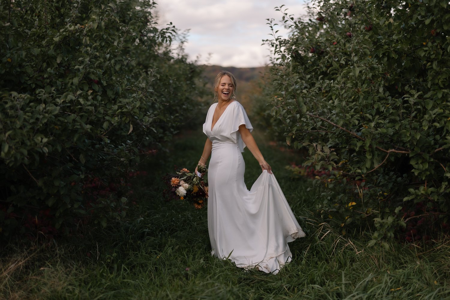Bride laughs while holding her dress in rows of apple trees.