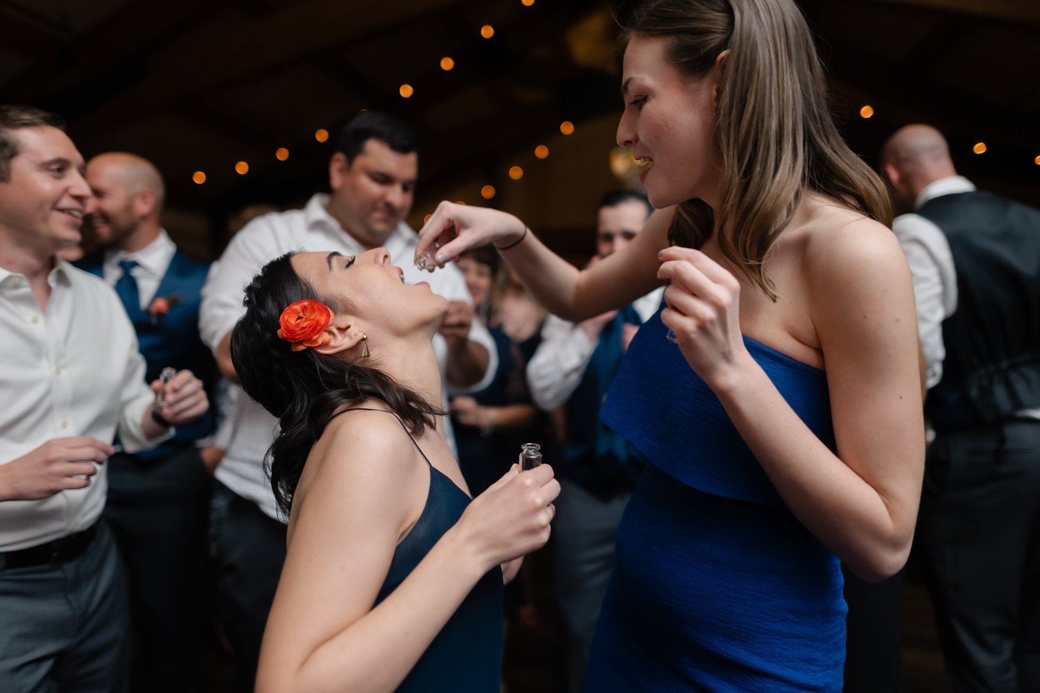 Girl in blue dress pours a shot of liquor into another girls mouth on wedding dance floor.