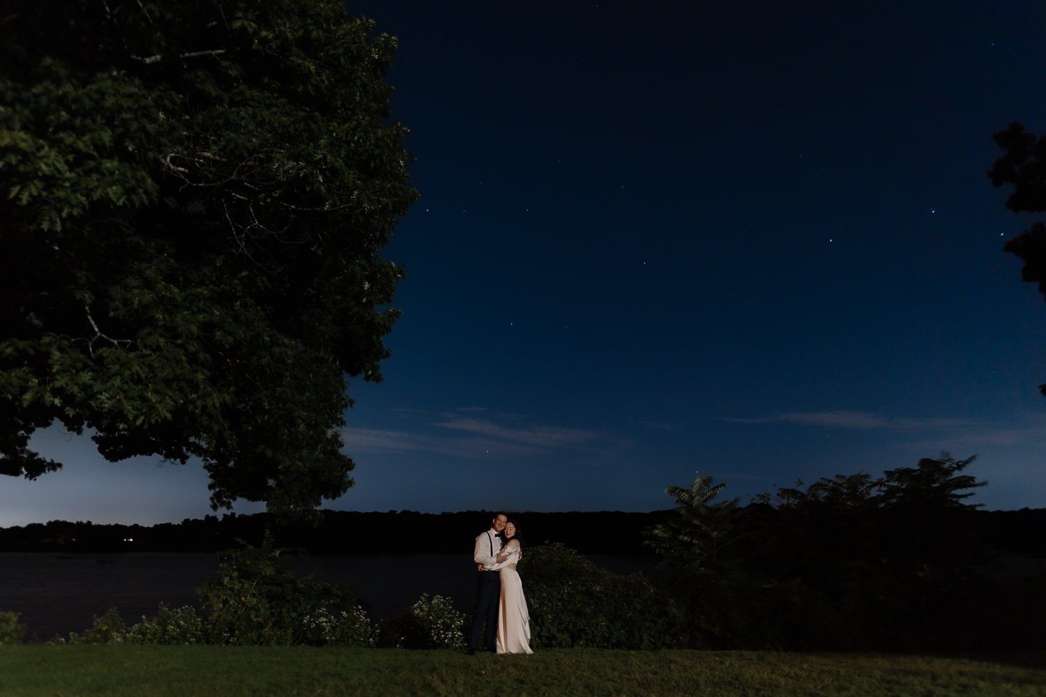 Married couple stands at rivers edge with blue night sky and stars behind them.