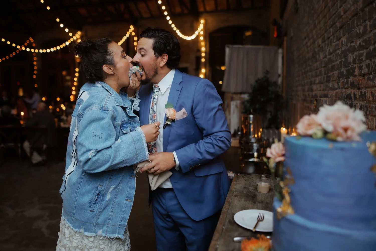 A woman in a denim jacket and a woman in a suit sharing a piece of cake at a wedding reception. The bride and groom are dressed in wedding attire and are in a decorated rustic venue with string lights. There is a large blue wedding cake with flowers on a table in the foreground.