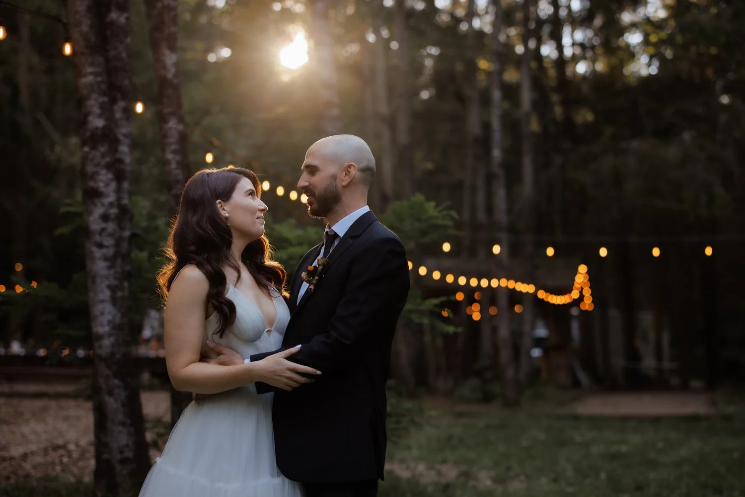 A couple in wedding attire holding each other and gazing into each other's eyes outdoors at sunset, with string lights hanging in the background in a wooded area.