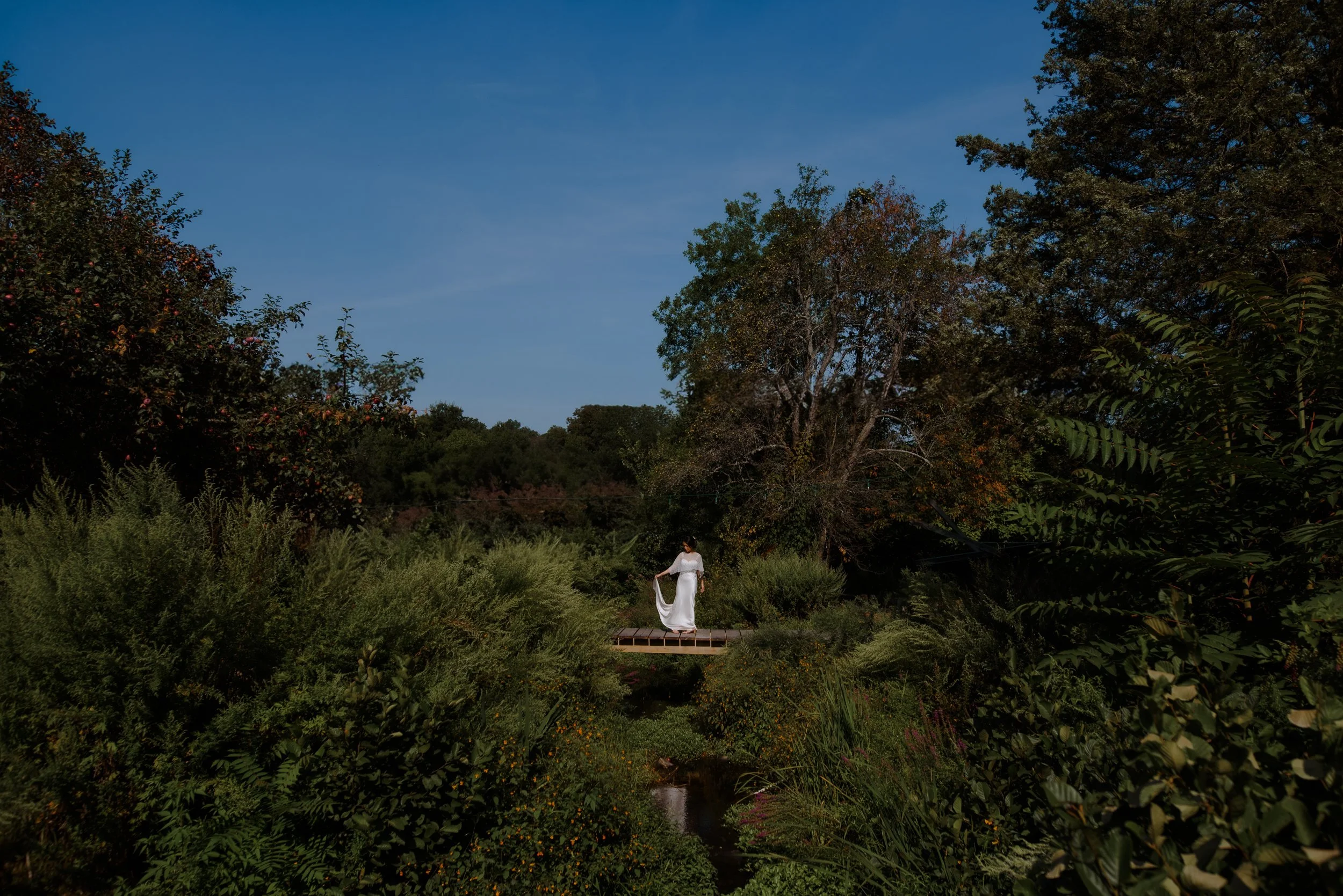 Bride crossing a small wooden bridge surrounded by dense summer greenery.
