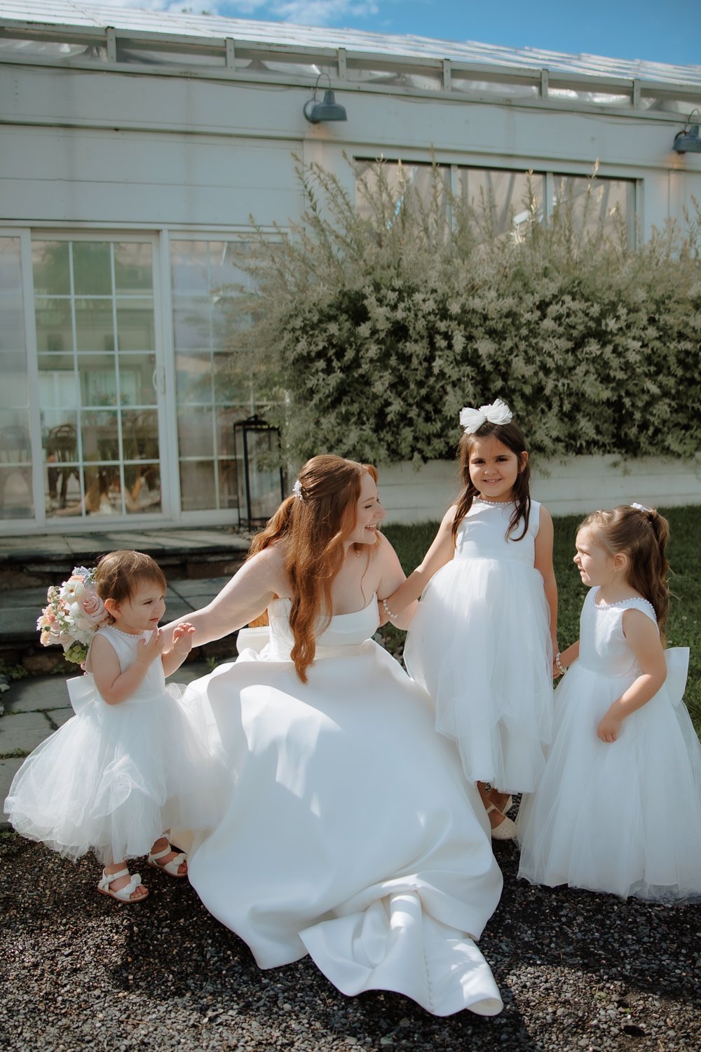 Bride laughing with three flower girls in white dresses in a garden setting.