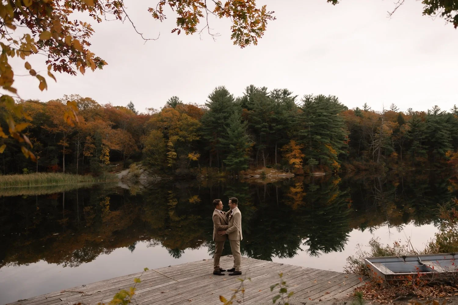 Two groom stand on dock with Autumn trees reflecting in the lake behind them.