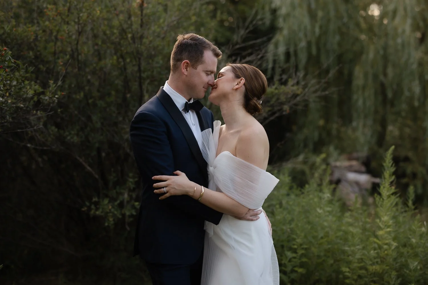 A newlywed couple shares an intimate moment outdoors, with their noses touching and eyes closed, surrounded by greenery.