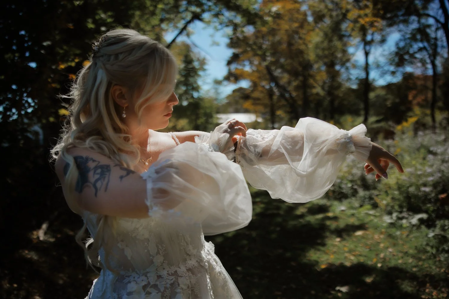 A woman with long blonde hair in a wedding dress with lace and ruffled sleeves, standing outdoors in a wooded area with sunlight filtering through trees, striking a contemplative pose.