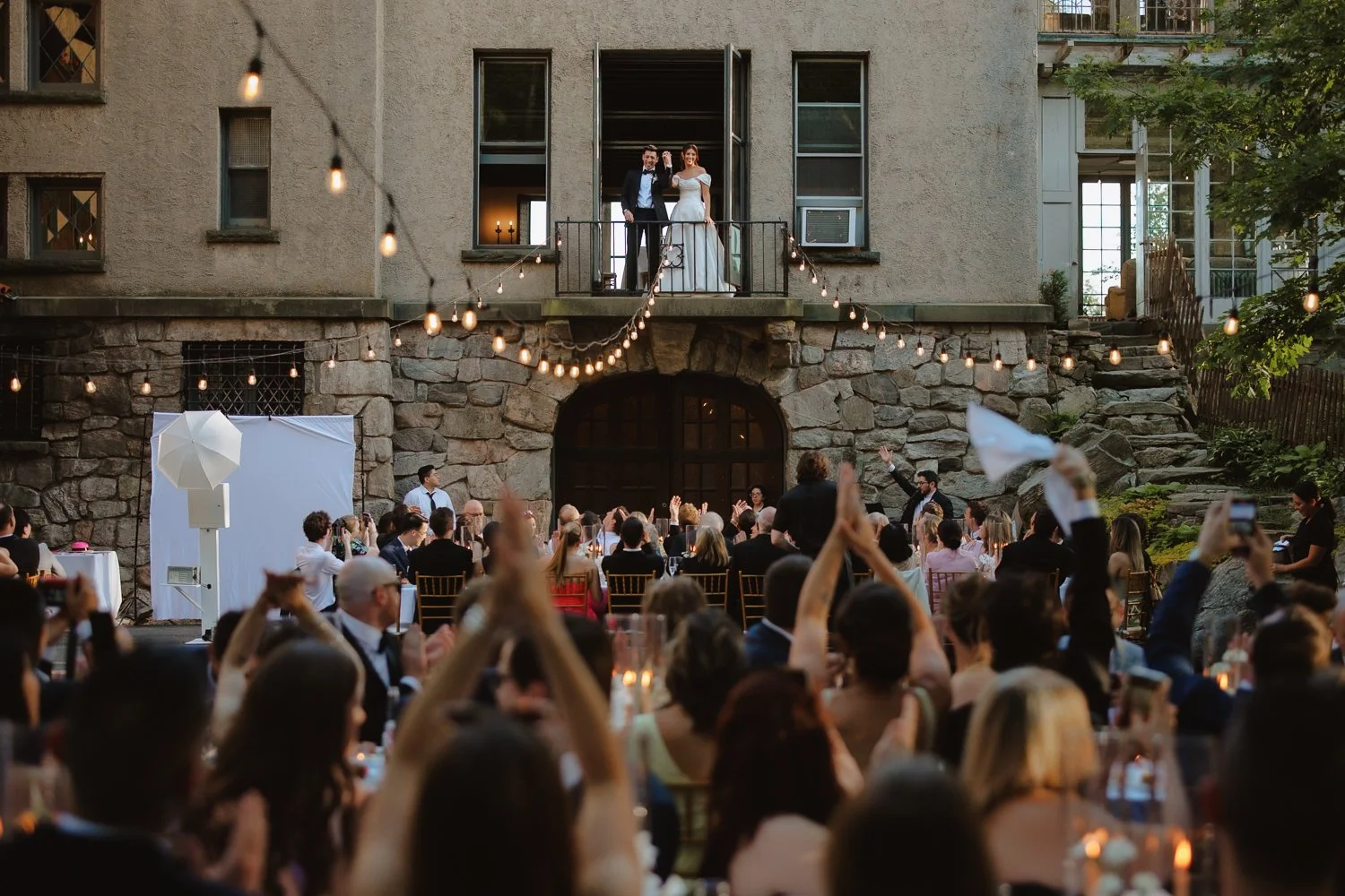 Outdoor wedding reception with a couple on balcony, guests seated, string lights overhead.