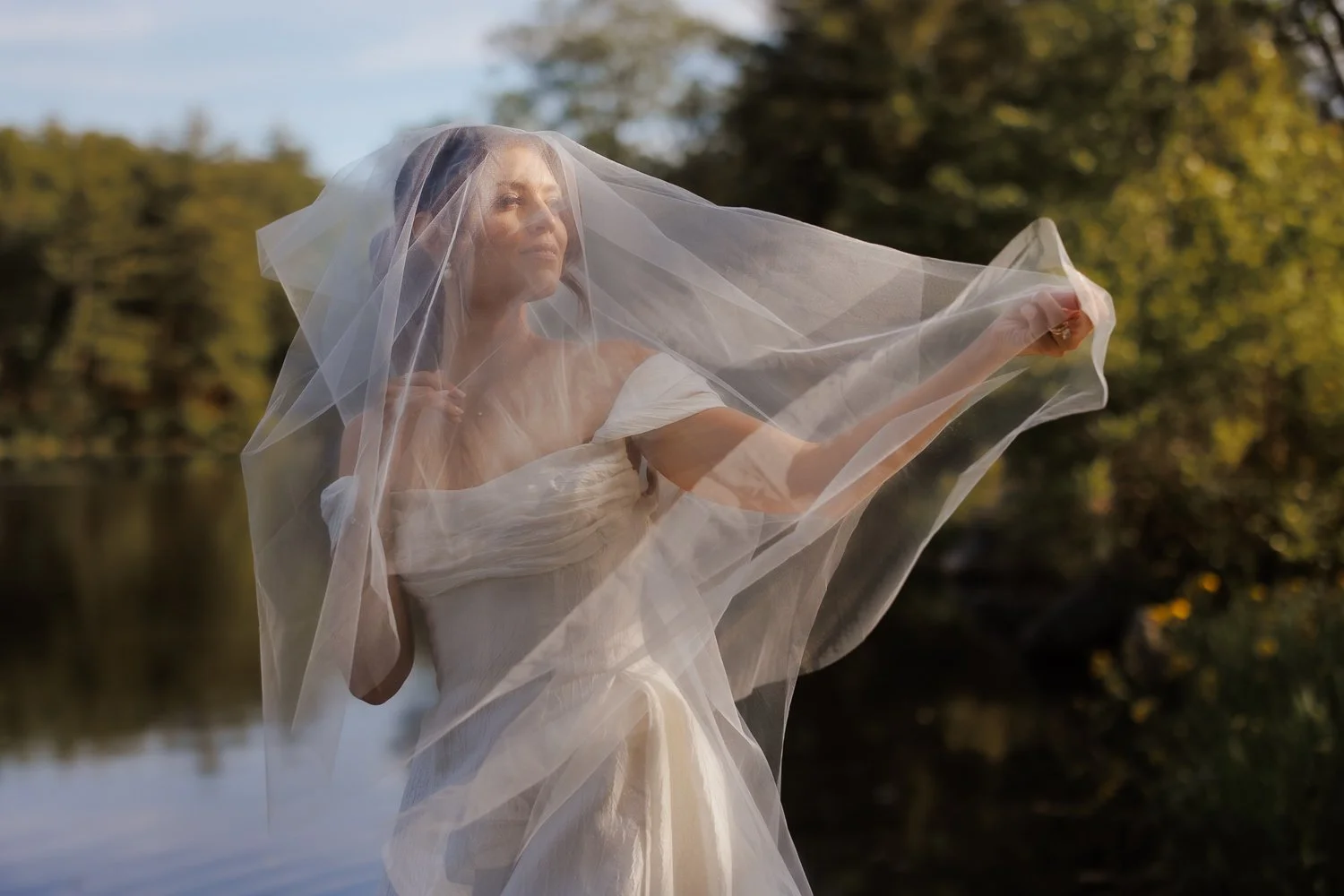 A woman in a wedding dress holding a veil outdoors by a body of water, with trees and a cloudy sky in the background.