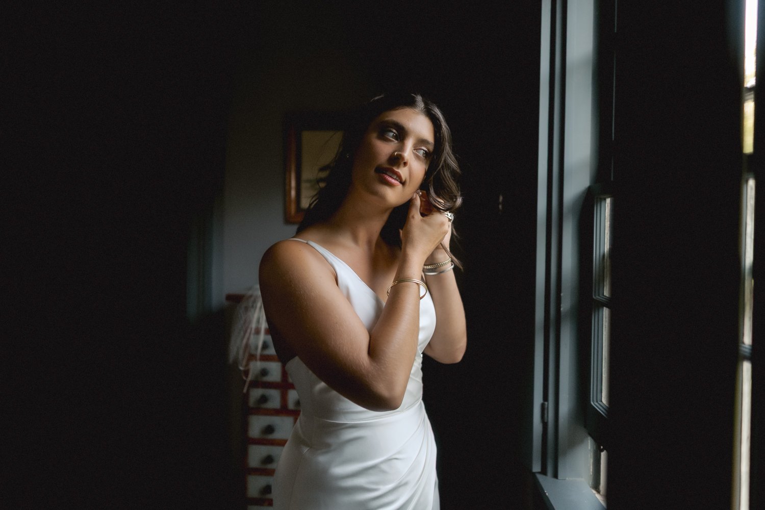 Bride puts on her earrings in moody window light at Catskills wedding.