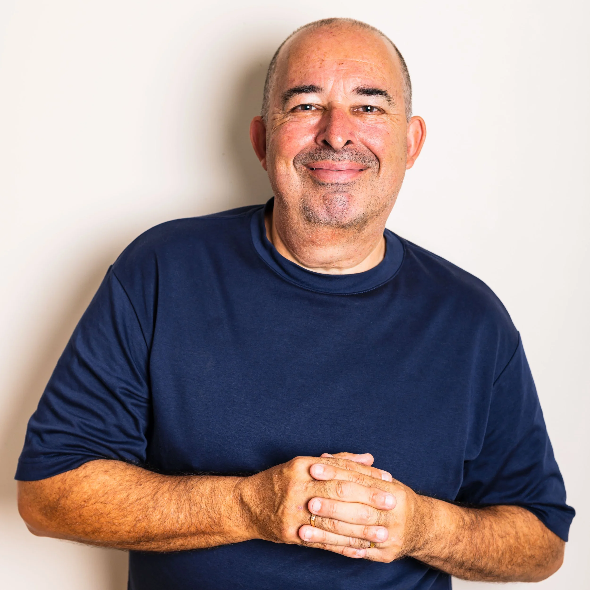 Close-up of a middle-aged man with short gray hair, holding his hands, wearing a dark blue shirt, standing against a plain light background.
