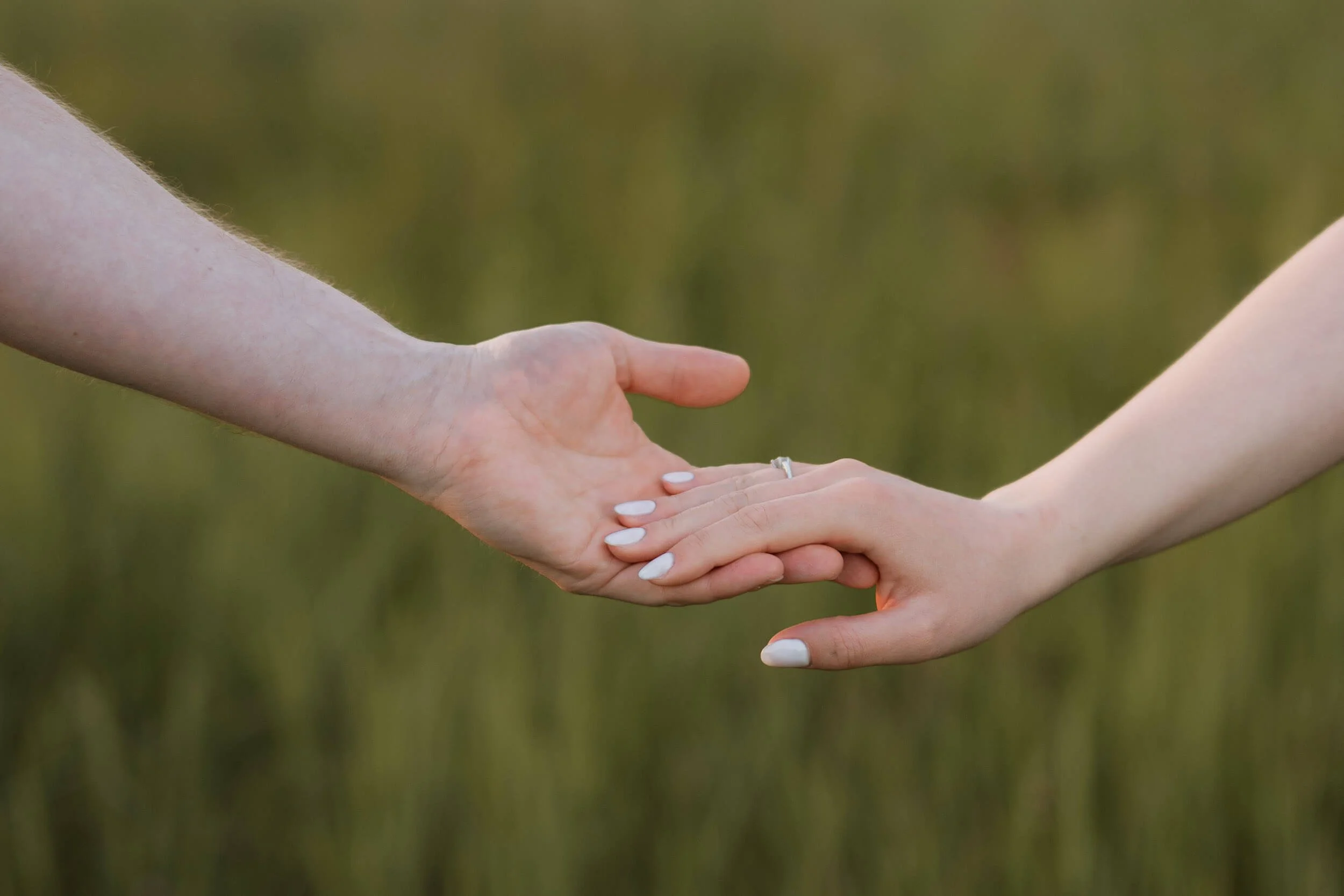 Two hands reaching towards each other, one with a wedding ring, against a blurred green background.