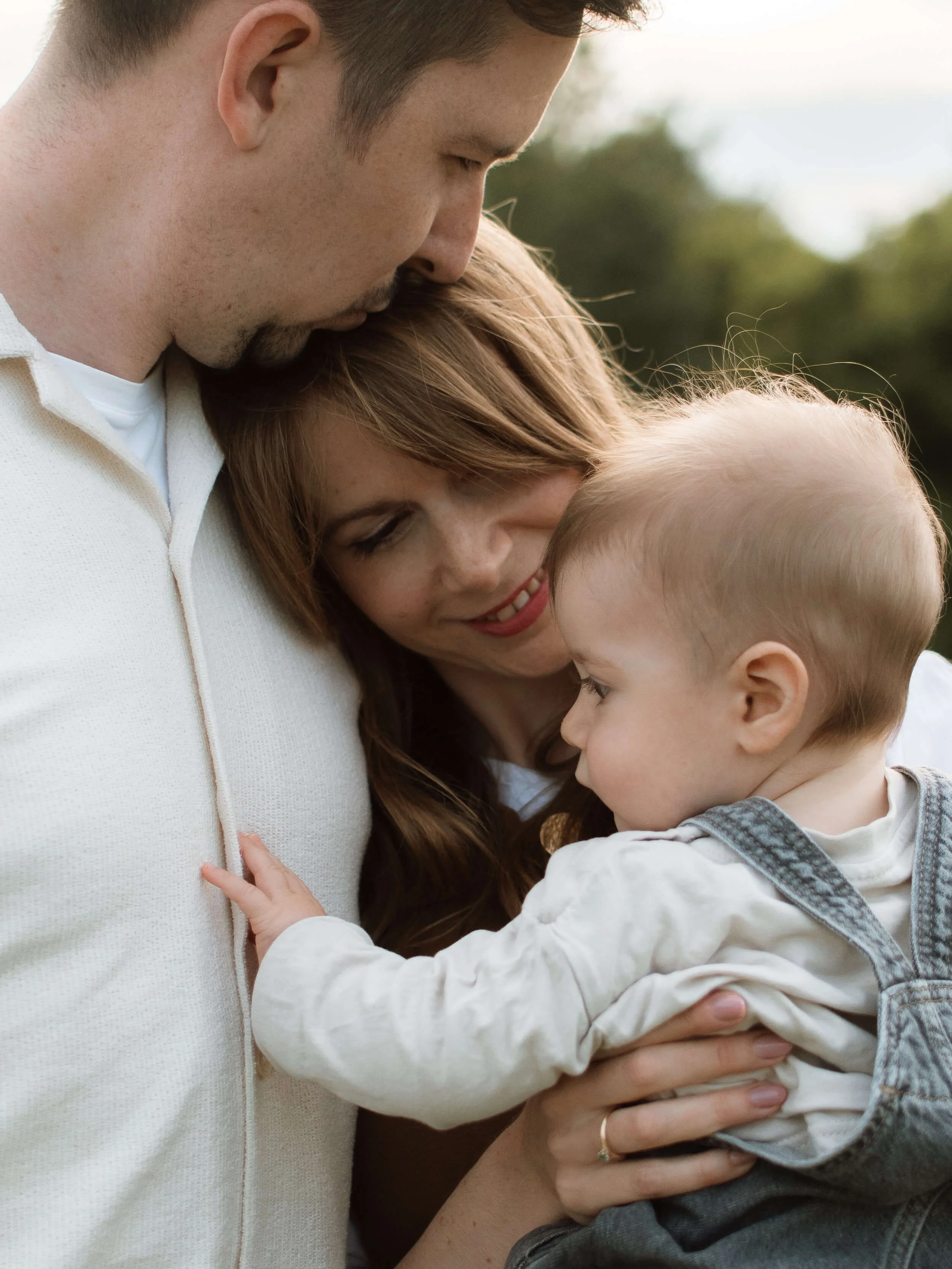 A man, woman, and young child are embracing outdoors, with their eyes closed and heads touching, expressing love and affection.