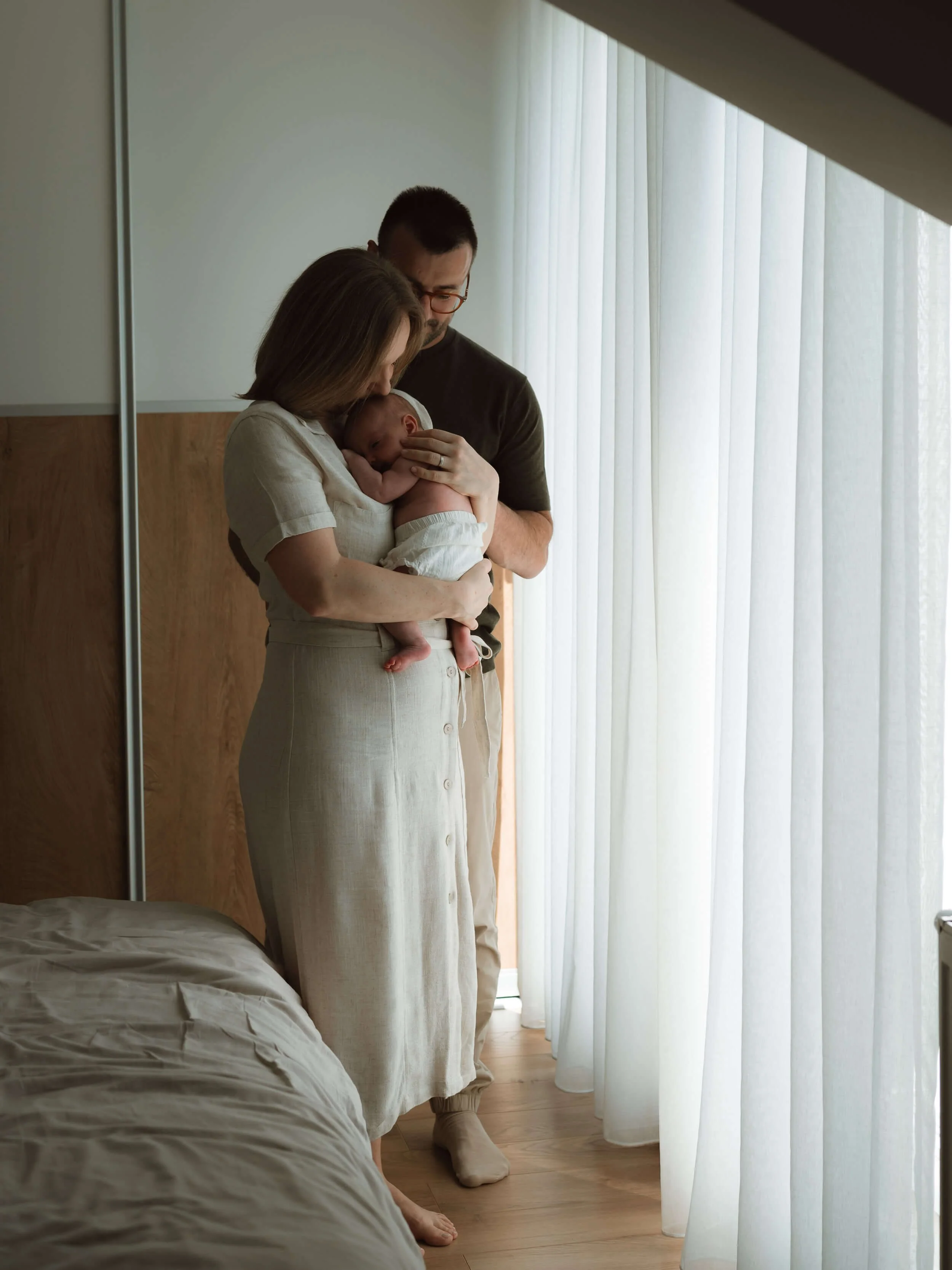 Two adults, a man and a woman, stand close together by a window with white sheer curtains. The woman holds a newborn baby, pressing it gently against her chest, while the man looks down lovingly. The scene is calm and tender, set in a room with wooden floors and a bed partially visible in the foreground.