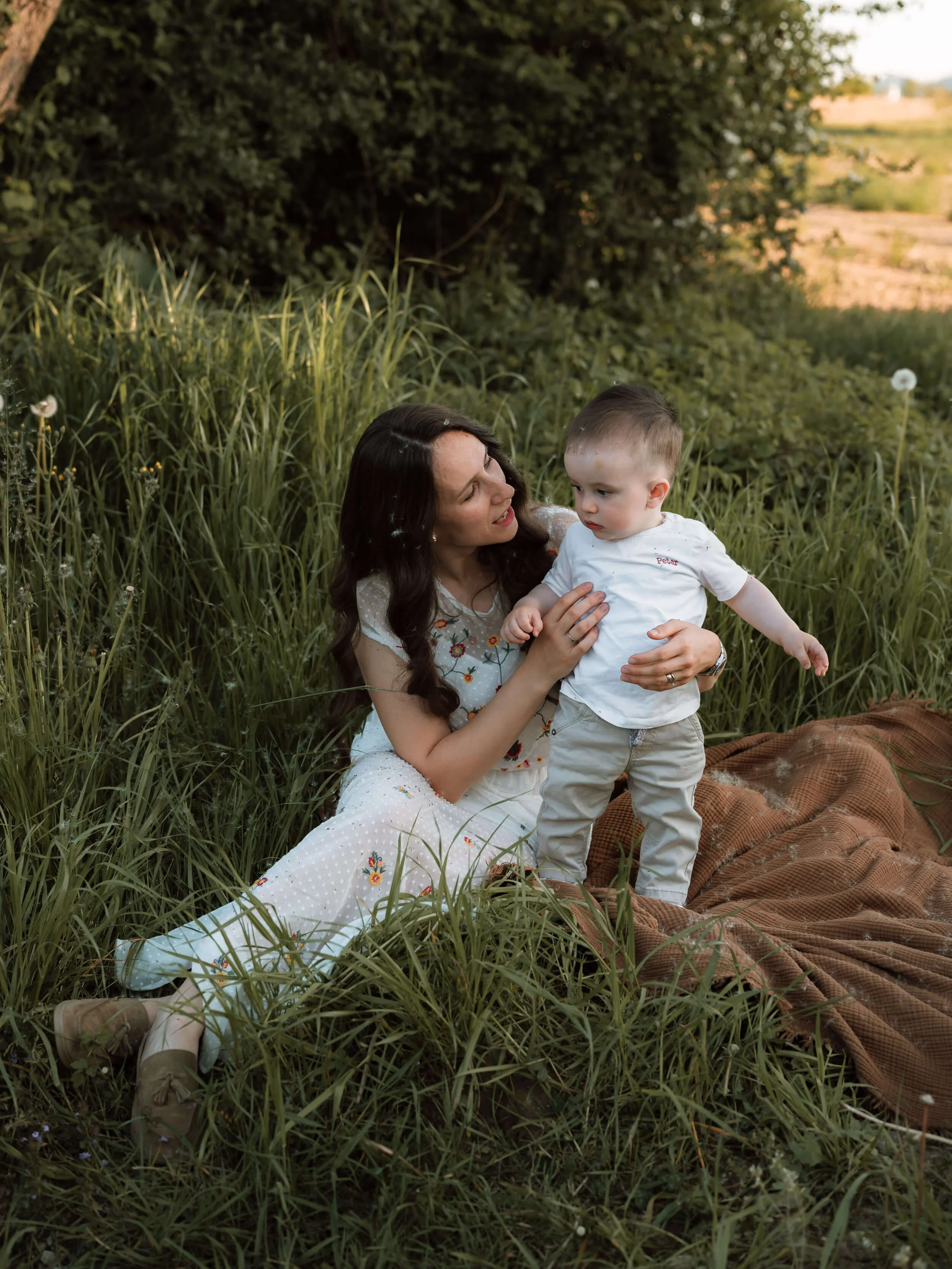 A woman with long dark hair and a young boy with short hair sitting in tall grass with greenery and trees in the background.