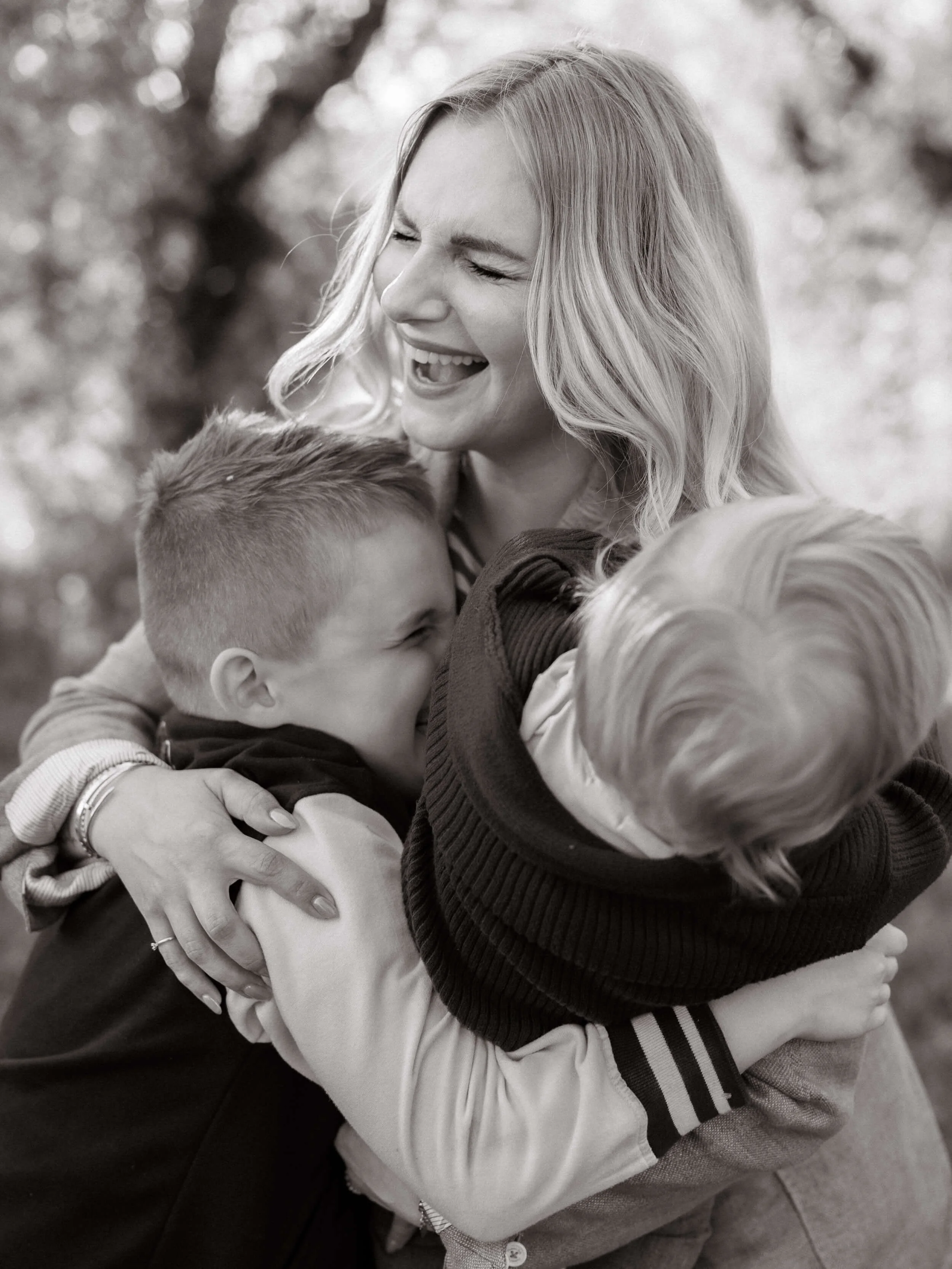 Black and white photo of a woman with blonde hair smiling and hugging two children, outdoors with blurred background.