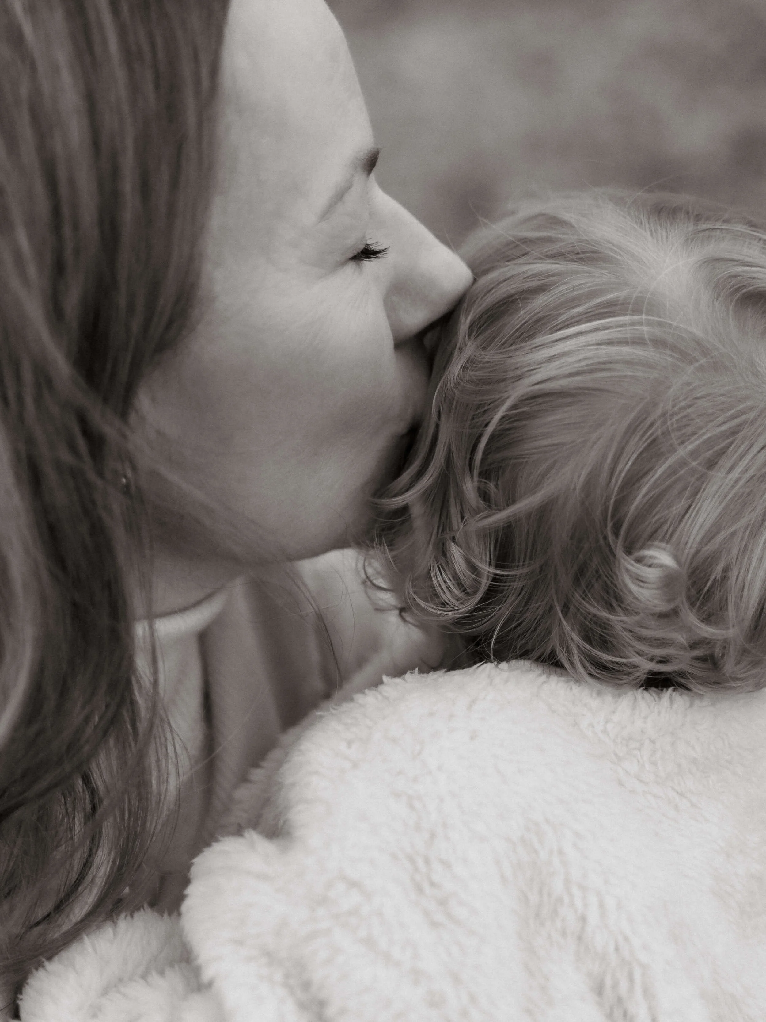 A woman with long dark hair and eyelashes nuzzles a blonde child's head with curly hair, both lying on a soft white surface.