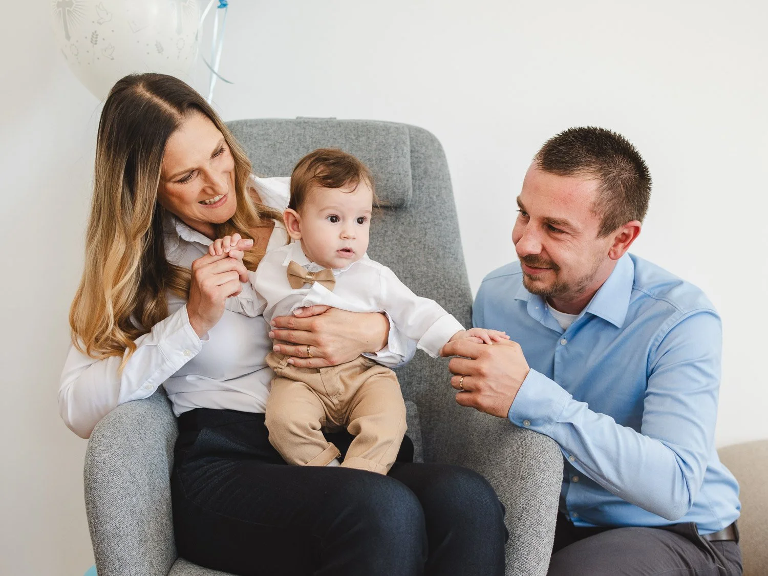 A family of three celebrating a special occasion with the mother holding a baby boy dressed in a white shirt and beige pants, and the father sitting nearby, all smiling and engaging with each other.