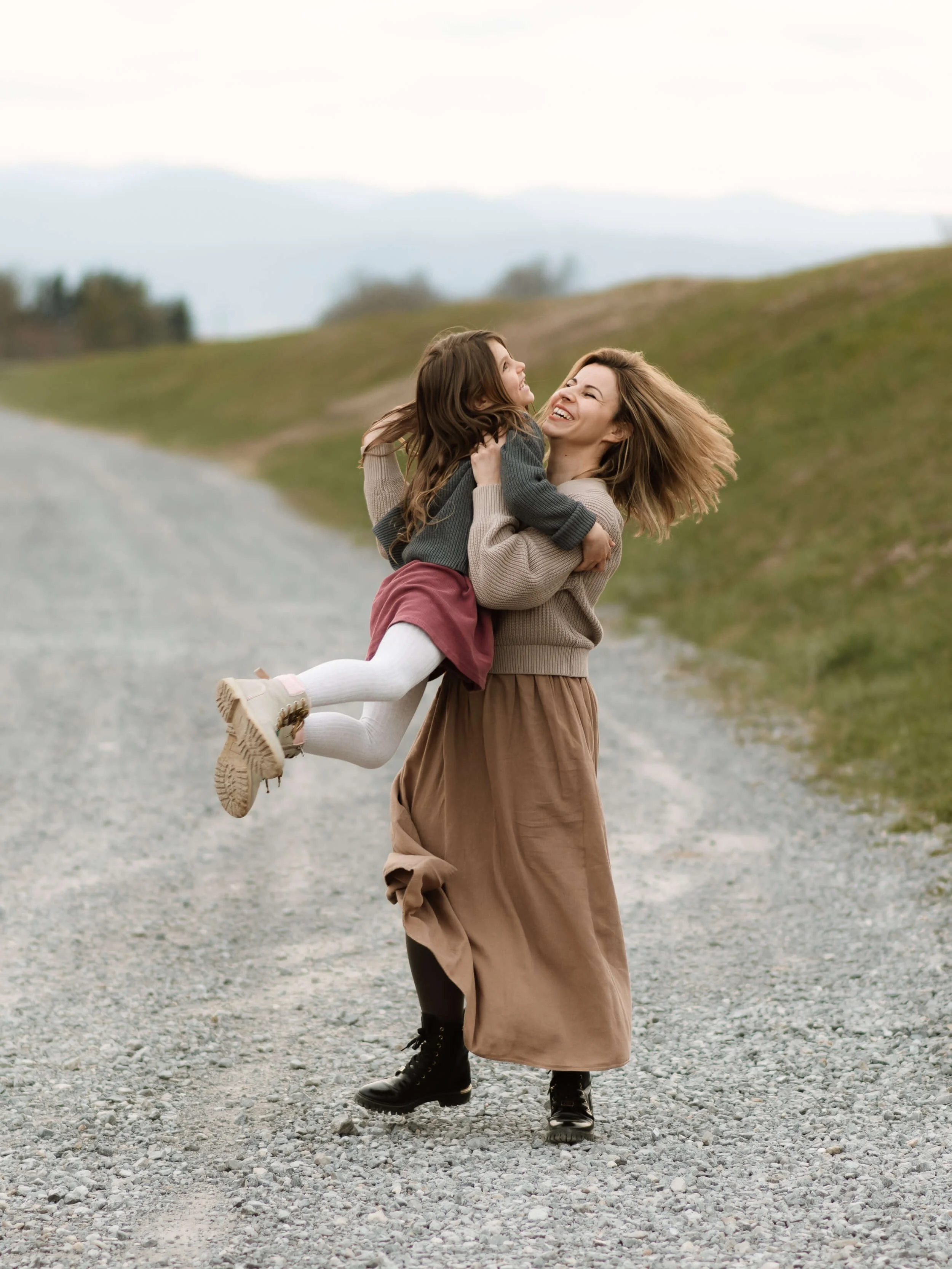 A woman with long hair wearing a long skirt, black boots, and a beige sweater lifts and holds a young girl with brown hair, white tights, pink shoes, and a gray and beige sweater in a scenic outdoor setting with grassy hills and cloudy sky.