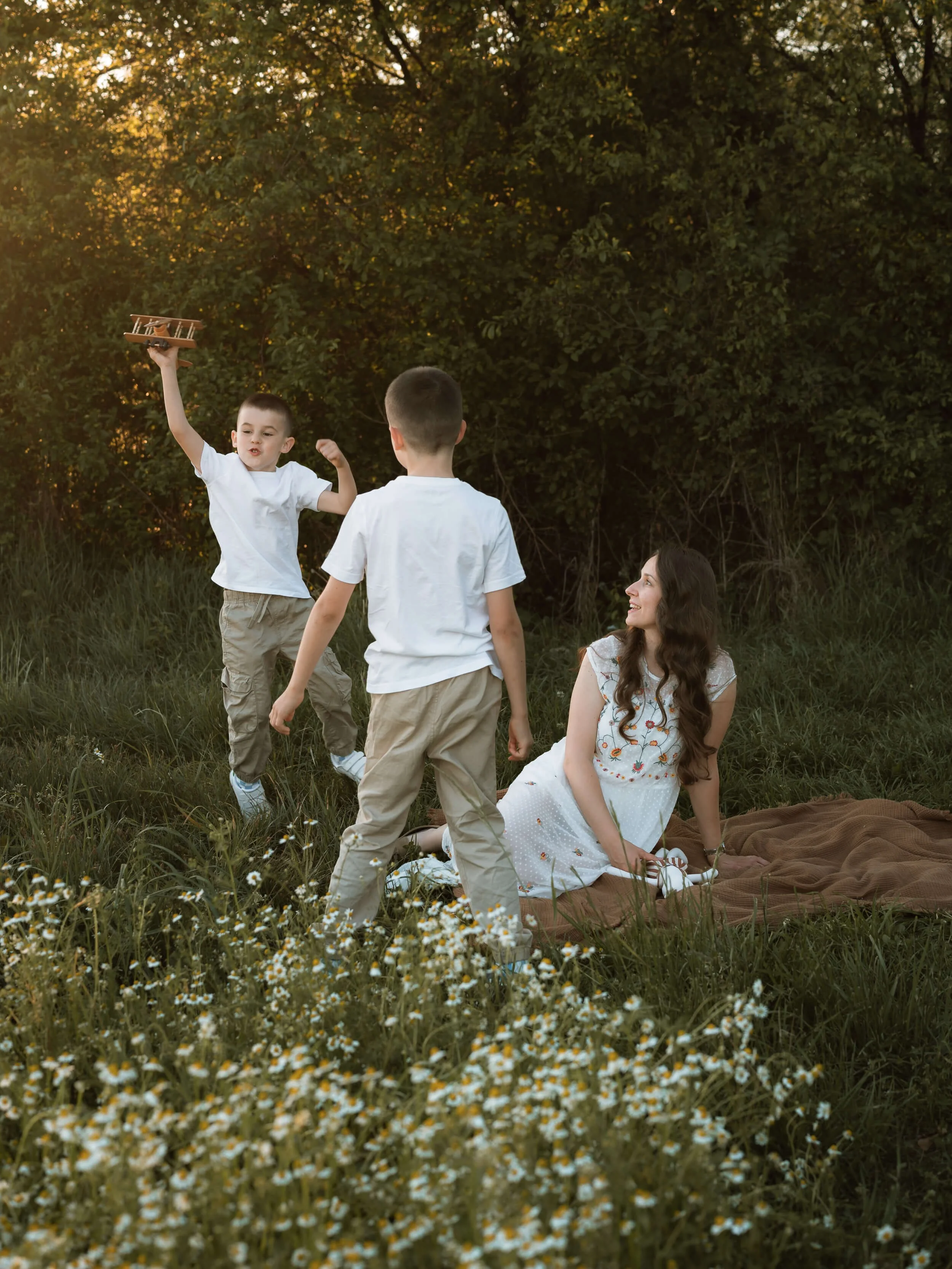 A woman sitting on a blanket in a grassy field with four children playing and jumping around her during sunset. One child holding a toy airplane in the air.