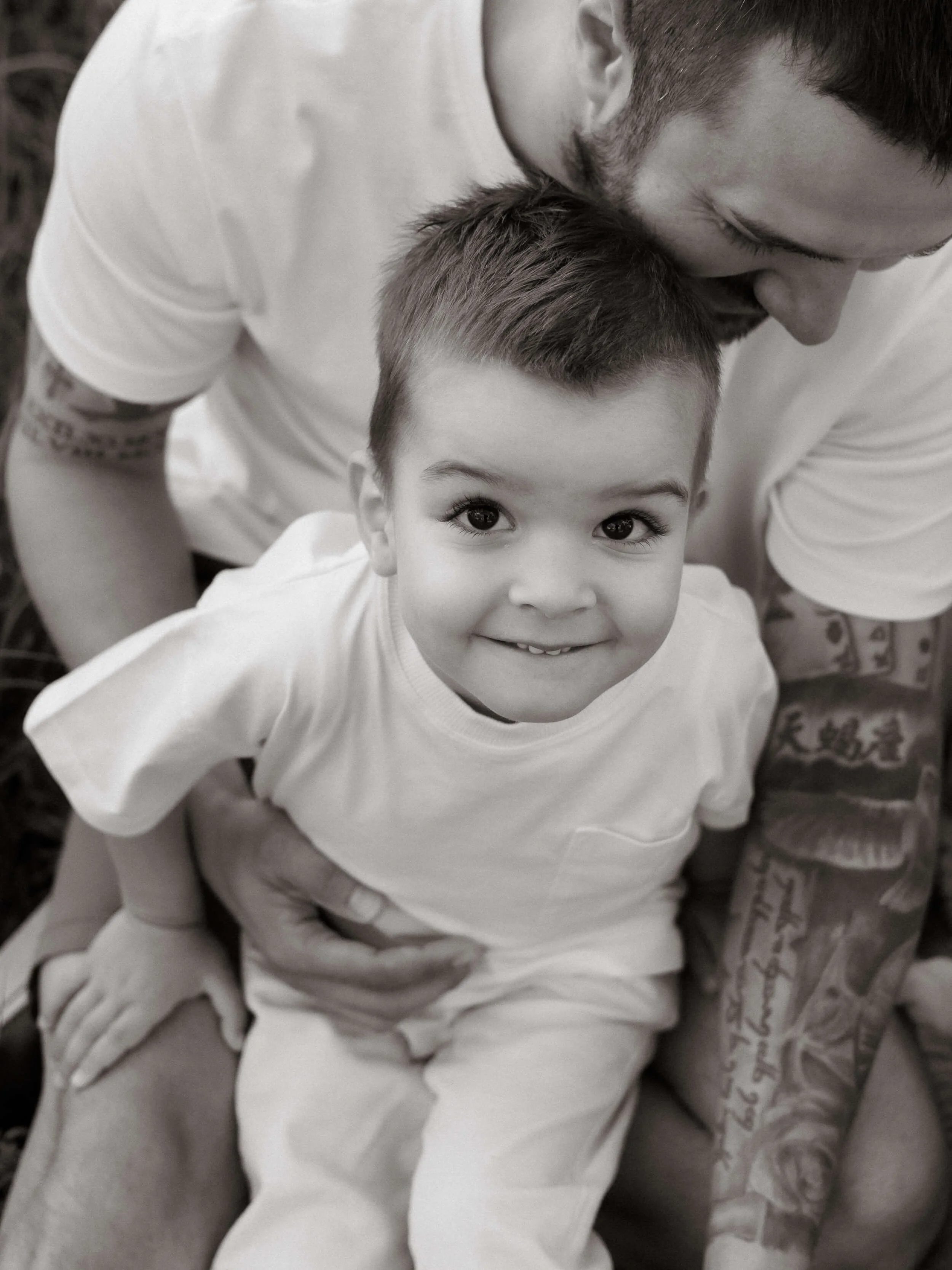A young boy with short hair sitting next to an adult male with facial hair, both smiling and looking up at the camera. The boy has large eyes and a cheerful expression.