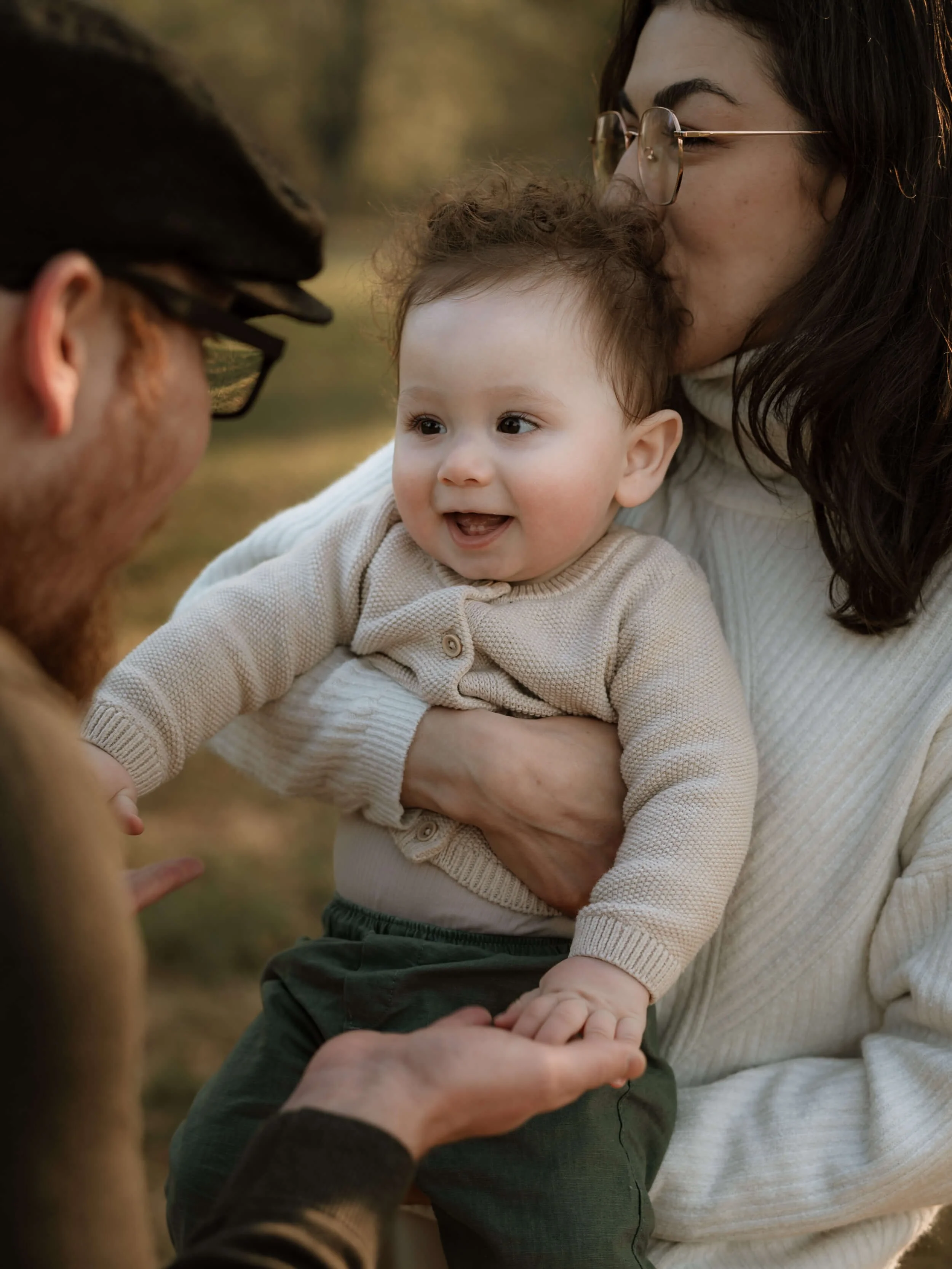 A woman with glasses kissing a child's forehead while a person in a hat and glasses holds the child's hand. The scene is outdoors with blurred trees in the background.