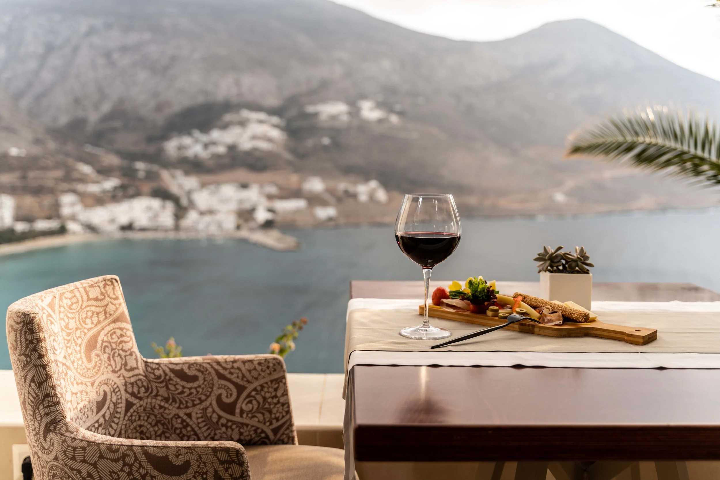 Sea-view terrace dining at Aegialis Hotel & Spa in Amorgos, Greece, with a glass of red wine and Mediterranean sharing platter.
