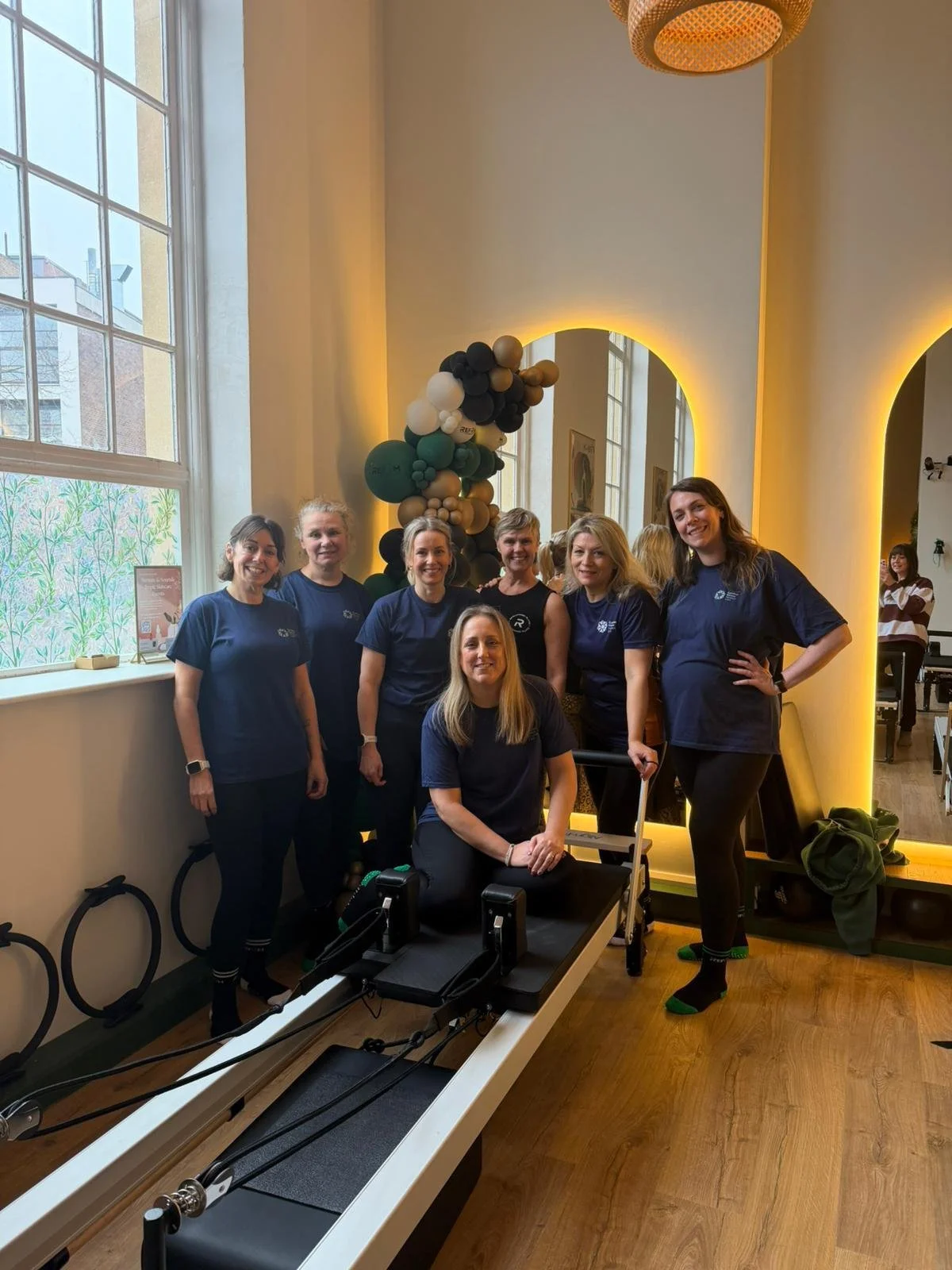 A group of eight women, some standing and one sitting, in a well-lit room with a large window, wood flooring, and decorative balloons, posing around a Pilates reformer fitness machine.
