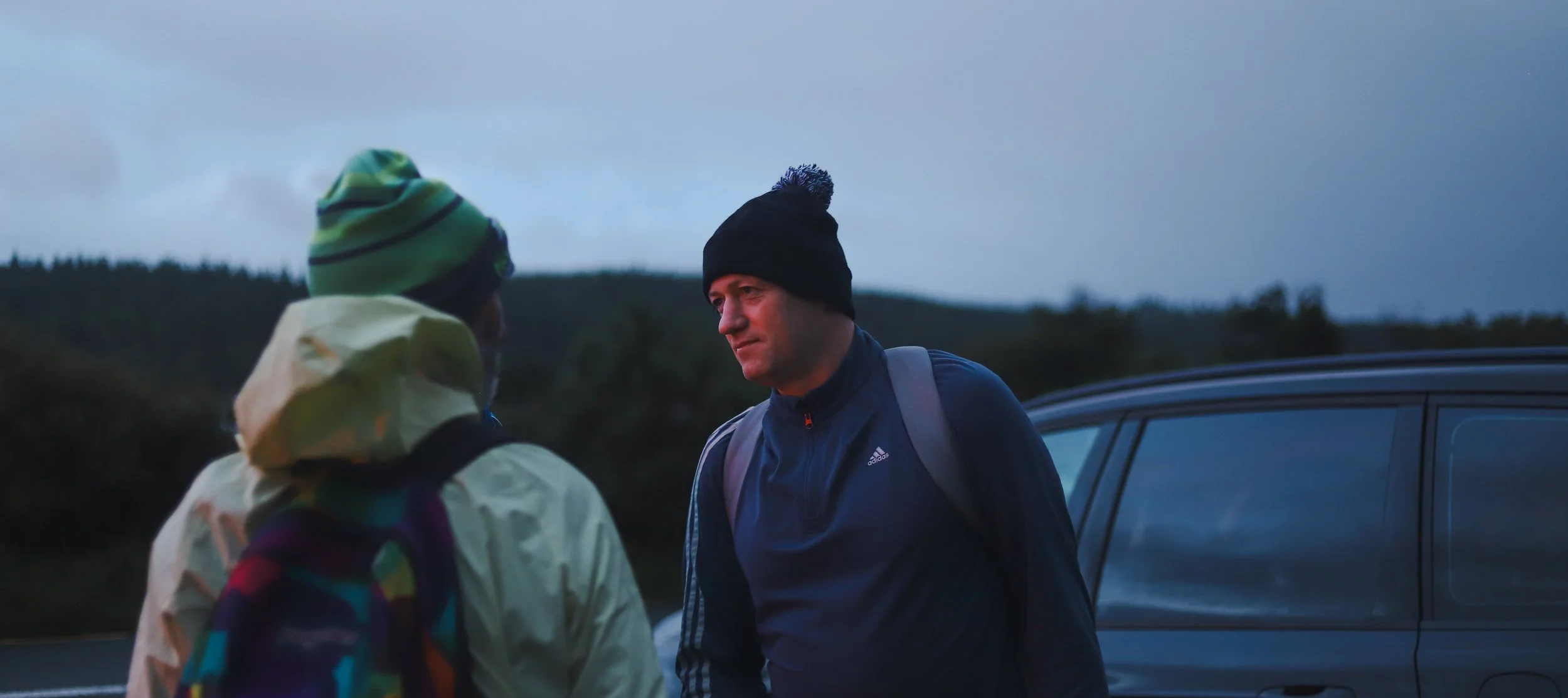 Two men in outdoor clothing and beanies talking beside a parked car in a natural setting at dusk.