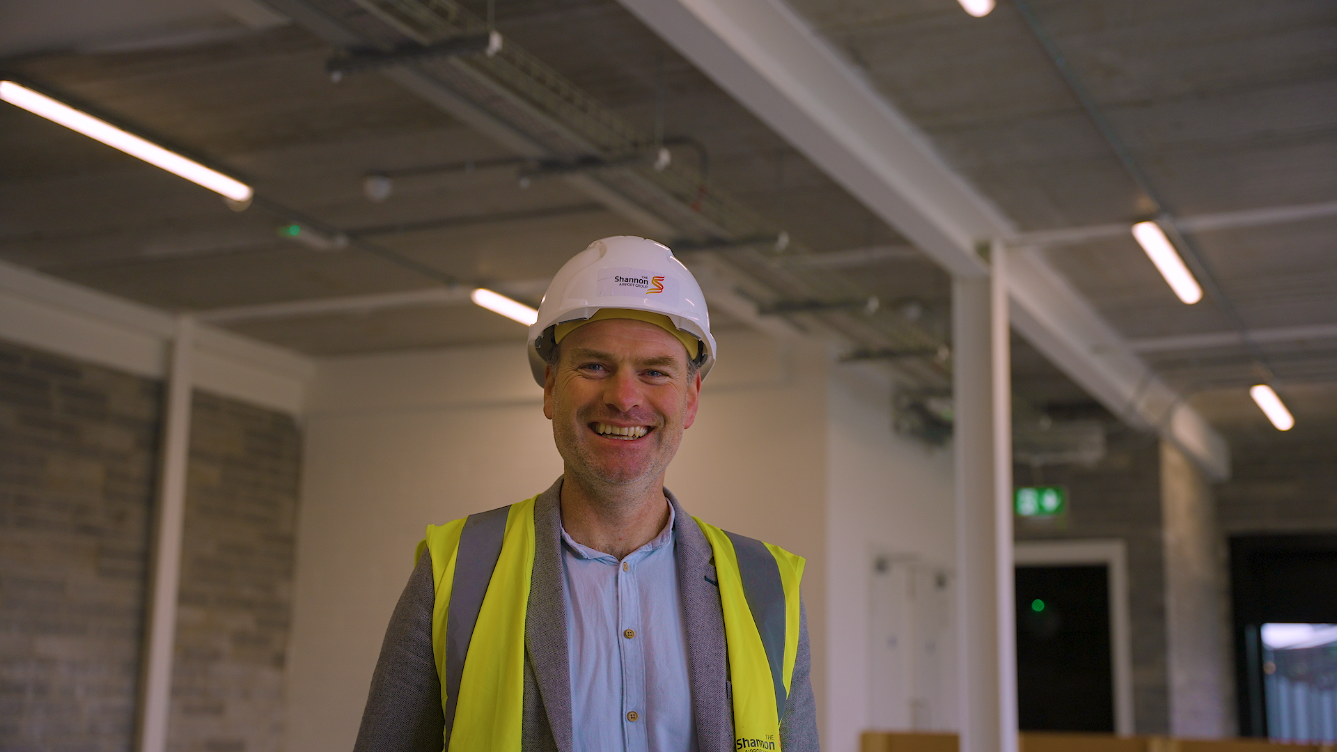 A man smiling in an indoor construction site wearing a white hard hat with 'Shannon' logo and a reflective yellow safety vest.