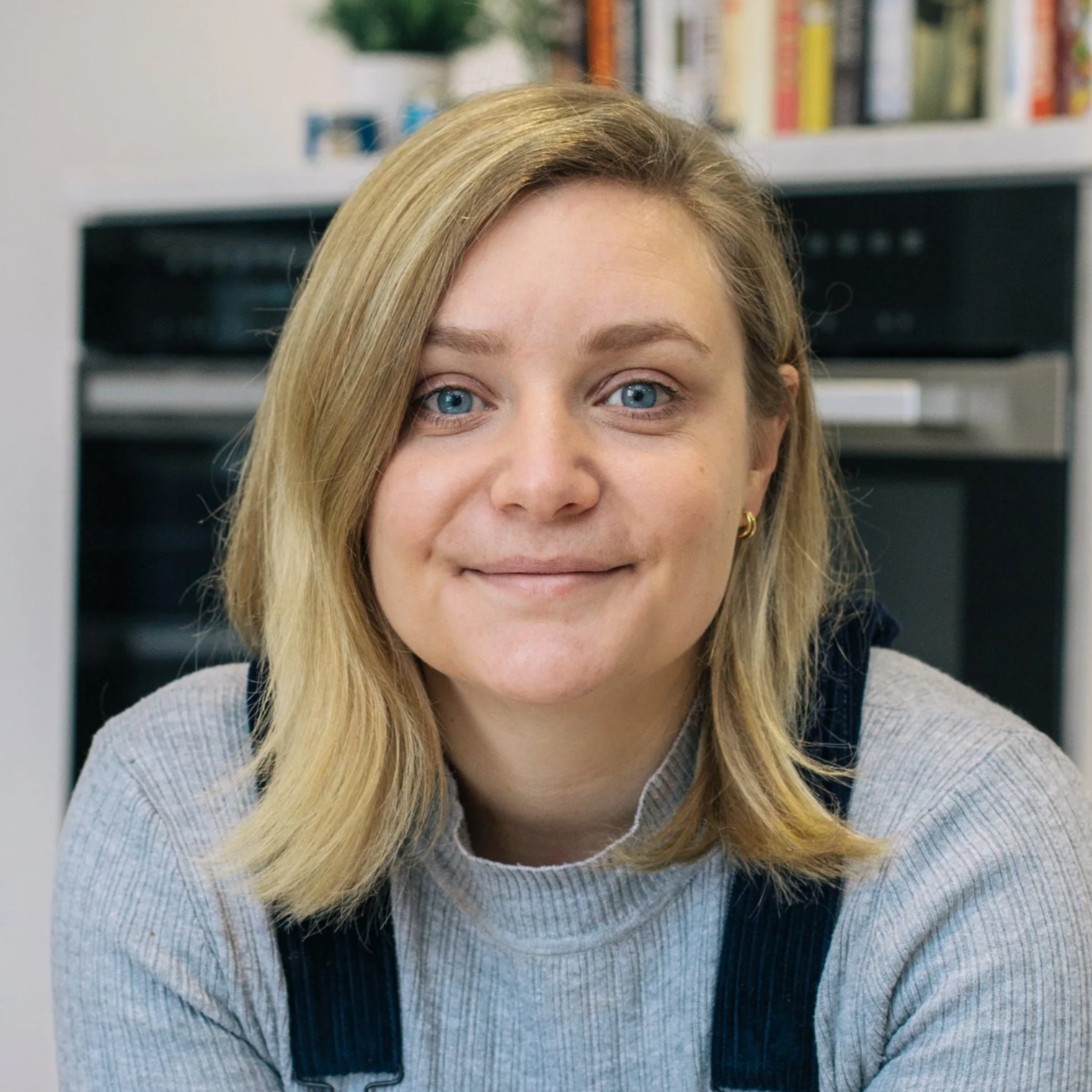 A young woman with blonde hair and blue eyes sitting at a kitchen table, smiling gently at the camera, with a background of a white wall, a black oven, and a shelf with books and plants.