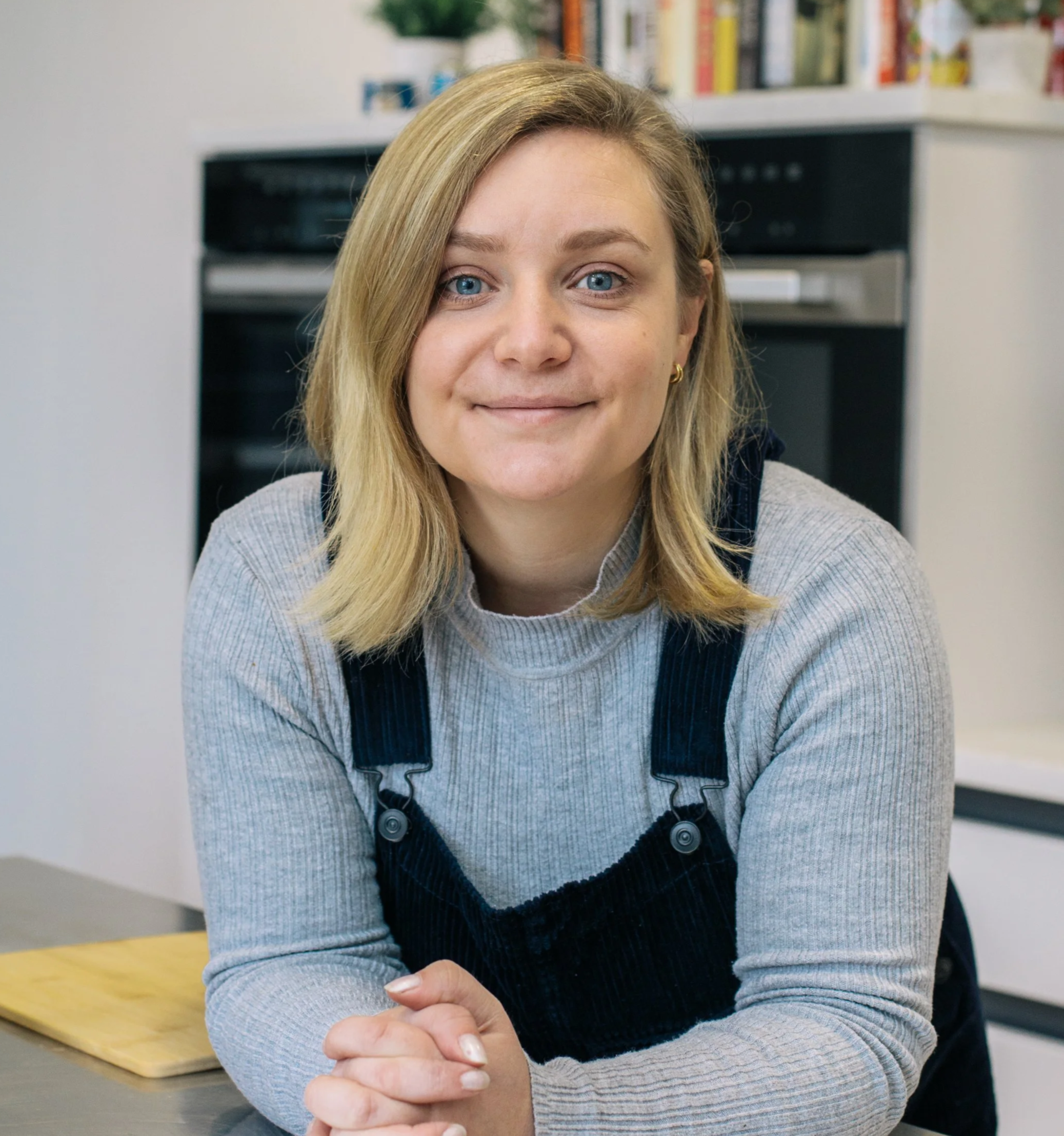 A young woman with blonde hair and blue eyes sitting at a kitchen table, smiling gently at the camera, with a background of a white wall, a black oven, and a shelf with books and plants.